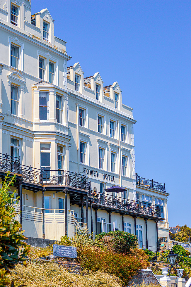 The image depicts a multi-story hotel named 'Fowey Hotel' with a white facade and numerous windows. The hotel has balconies with wrought iron railings and is surrounded by some greenery and a clear blue sky in the background.