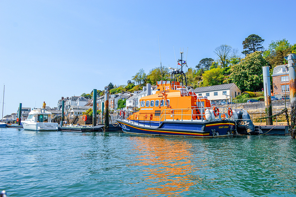 The image depicts a vibrant scene at a harbor. Dominating the foreground is a bright orange and blue lifeboat docked at a pier. The lifeboat is equipped with various safety gear, including life rings and a mast with a flag. In the background, there are several other boats, including a white yacht. The harbor is lined with buildings and lush greenery, creating a picturesque coastal setting under a clear blue sky.