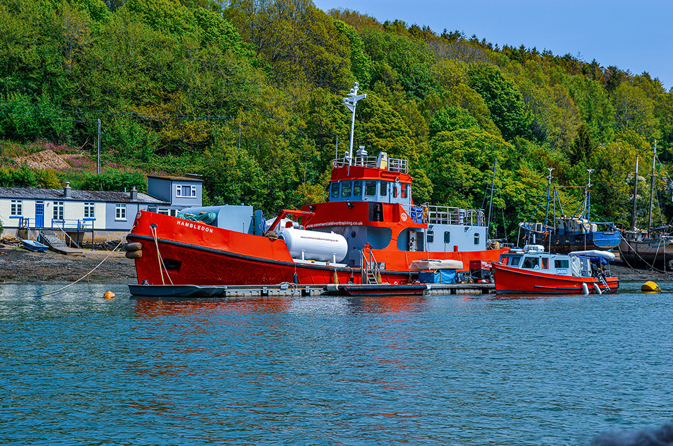 The image shows a harbor scene with several boats docked. The most prominent boat is a large red vessel named 'Hambleton,' which appears to be a tugboat or a similar type of working boat. The background features a hill covered with lush green trees and some small buildings near the water's edge. The water is calm, reflecting the boats and the surrounding scenery.