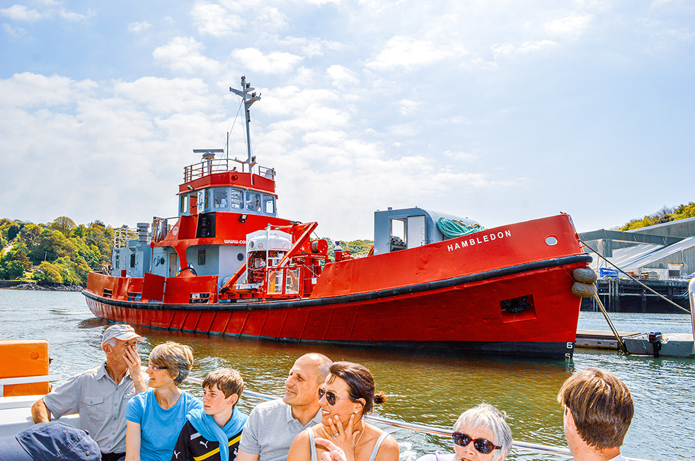 The image shows a red tugboat named 'Hambledon' docked at a pier with several people on a smaller boat in the foreground observing it. The scene is set on a calm body of water with a backdrop of trees and a partly cloudy sky. The people on the smaller boat appear to be tourists or visitors, enjoying the view of the tugboat.