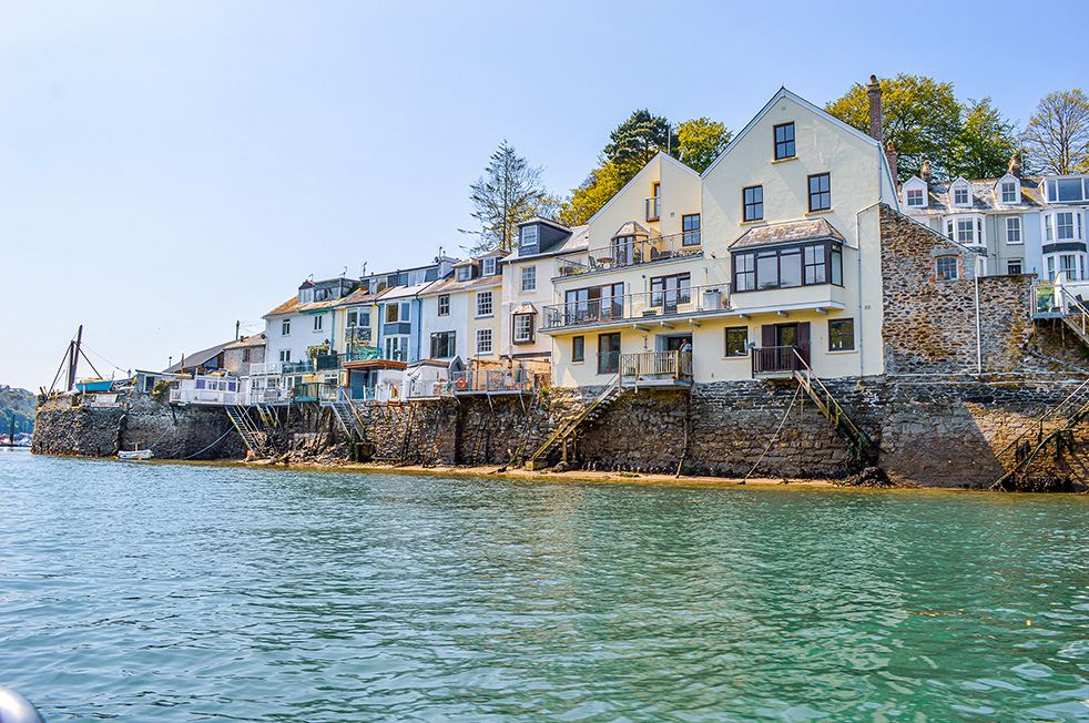 The image depicts a picturesque waterfront scene with a row of buildings situated along the edge of a body of water. The buildings vary in architectural style, with some having modern balconies and others featuring traditional stone facades. The water is calm, reflecting the clear blue sky above. The overall atmosphere is serene and inviting, suggesting a coastal or lakeside location that could be a popular spot for residents and visitors alike.