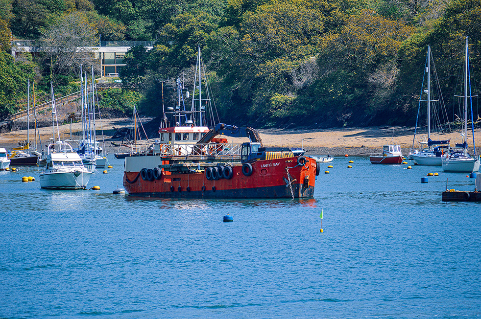 The image depicts a coastal scene with several boats anchored in a bay. The central focus is a red and black boat named 'Lantic Bay,' which appears to be a fishing vessel. Surrounding it are various sailboats and yachts moored in the calm, blue water. In the background, there is a sandy beach, a rocky shoreline, and a hillside covered with trees and a house. The overall atmosphere is serene and picturesque, typical of a peaceful harbor setting.