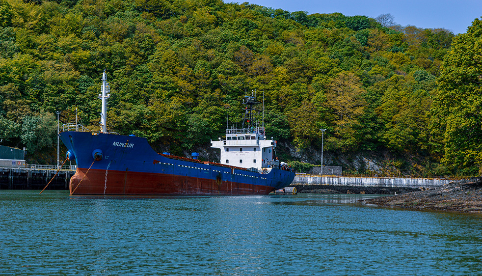 The image shows a large cargo ship named 'MUNZUR' docked at a port. The ship is blue and red, and it is moored next to a concrete pier. The background features a lush, green forested hillside, and the water in the foreground is calm. The scene appears to be in a serene, natural setting with no other vessels or significant activity visible.