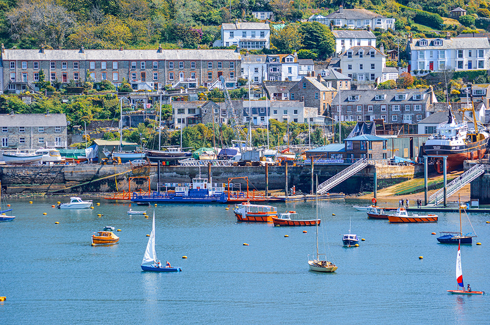 The image depicts a picturesque harbor scene with various boats anchored in the water. In the background, there are several multi-story buildings, likely residential houses and possibly some commercial establishments, situated on a hillside. The harbor is equipped with docks and moorings, and there are buoys marking different areas in the water. The overall setting appears to be a coastal town or village with a serene and scenic environment.