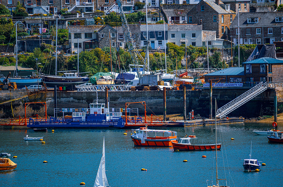 The image depicts a vibrant harbor scene with various boats and ships docked. In the foreground, there are smaller boats moored in the water, while a larger blue ship is docked at a pier. The background features a hillside with numerous houses and buildings, creating a picturesque coastal town atmosphere. The harbor is bustling with activity, and the overall scene is colorful and lively.