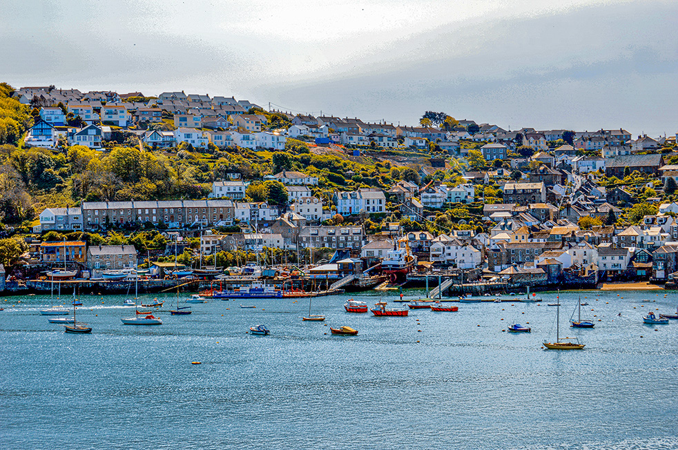The image depicts a picturesque coastal town with a harbor filled with boats. The town is built on a hillside, with houses and buildings cascading down to the waterfront. The scene is vibrant, with a mix of residential and commercial structures, and the water is calm, reflecting the boats and the town.