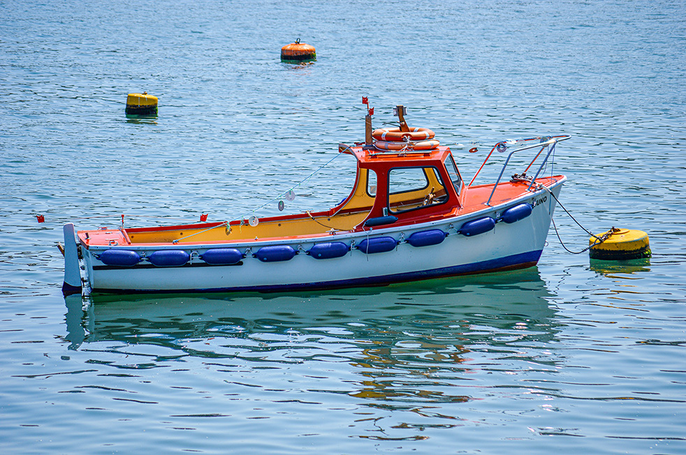 The image shows a small boat floating on calm water. The boat is primarily white with blue and orange accents. It has a cabin with a red roof and several life preservers are visible on top of the cabin. The boat is equipped with various fishing gear and is anchored near a yellow buoy. In the background, there are additional buoys marking the area.