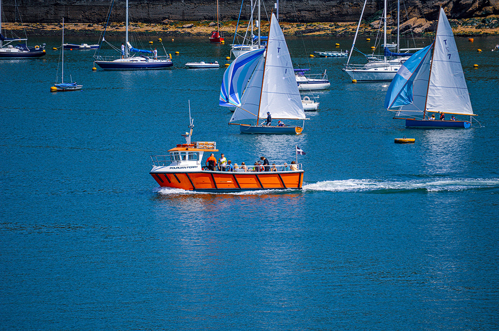The image depicts a vibrant harbor scene with various boats and yachts. In the foreground, there is an orange boat named 'Polruan Ferry' carrying passengers. Surrounding the ferry, several sailboats with white and blue sails are navigating the calm blue waters. The background shows a rocky shore with more boats anchored, creating a lively and bustling maritime atmosphere.