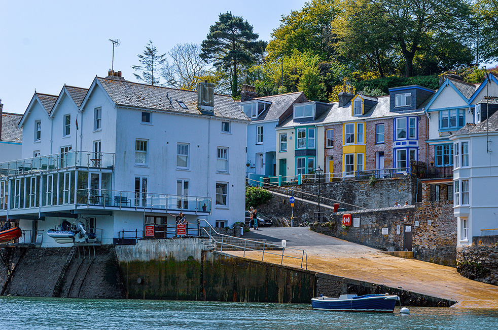The image depicts a picturesque waterfront scene with a row of colorful houses and buildings along the shore. The structures are predominantly white, with some featuring vibrant accents in blue, yellow, and green. The buildings have multiple stories, with some having balconies and large windows facing the water. A small boat is docked in the water in front of the buildings, and a stone wall and steps lead up from the waterfront to the buildings. The background includes lush green trees and a clear blue sky, adding to the serene and charming atmosphere of the scene.