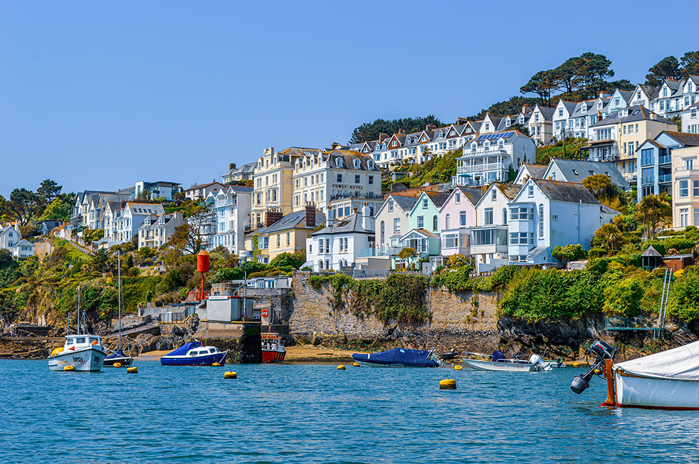 The image depicts a picturesque coastal town with a variety of colorful houses and buildings situated on a hillside overlooking a harbor. The harbor is dotted with several boats of different sizes and colors, anchored near the shore. The scene is vibrant and serene, with clear blue water and a bright, sunny sky. The town appears to be a charming and inviting place, likely a popular destination for tourists and visitors.
