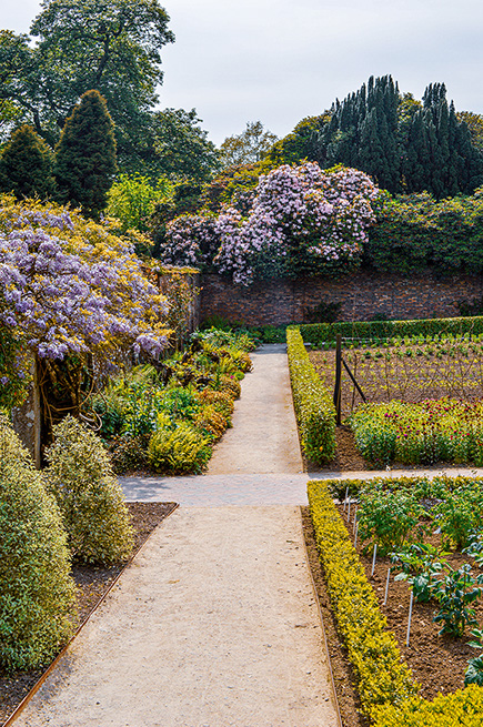 The image depicts a well-maintained garden with a central pathway leading through various sections of neatly arranged plants and flowers. The garden is bordered by a brick wall and lush greenery, showcasing a variety of colorful blooms and neatly trimmed hedges.