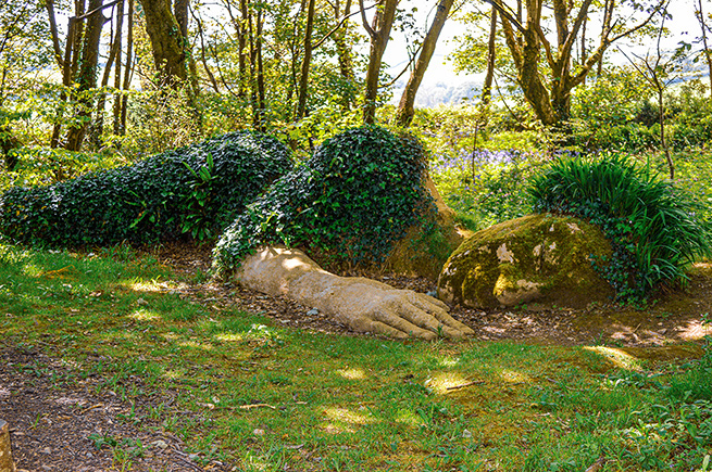 The image depicts a garden scene with a large, sculpted topiary of a sleeping dog. The topiary is crafted from green shrubbery and is situated on a grassy area surrounded by various plants and trees. The dog's form is detailed, showing its head resting on its paws, creating a serene and whimsical atmosphere.