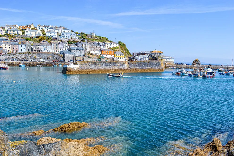 The image depicts a picturesque coastal town with a harbor. The town is built on a hillside, with houses stacked in rows, showcasing a variety of colors. The harbor is filled with boats, and the water is a clear, inviting blue. The foreground features rocky outcrops, adding to the scenic beauty of the location. The overall atmosphere is serene and idyllic, typical of a charming seaside village.