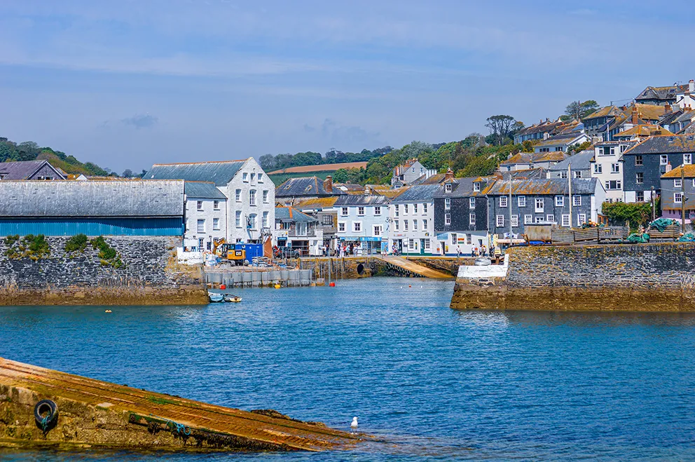 The image depicts a picturesque coastal town with a harbor. The town features a mix of white and colorful buildings along the waterfront, with a large white building prominently situated near the harbor entrance. The harbor is calm, with clear blue water, and is bordered by stone walls. In the background, there are hills with more houses and greenery, under a clear blue sky with some clouds. The overall scene is serene and charming, typical of a quaint seaside village.