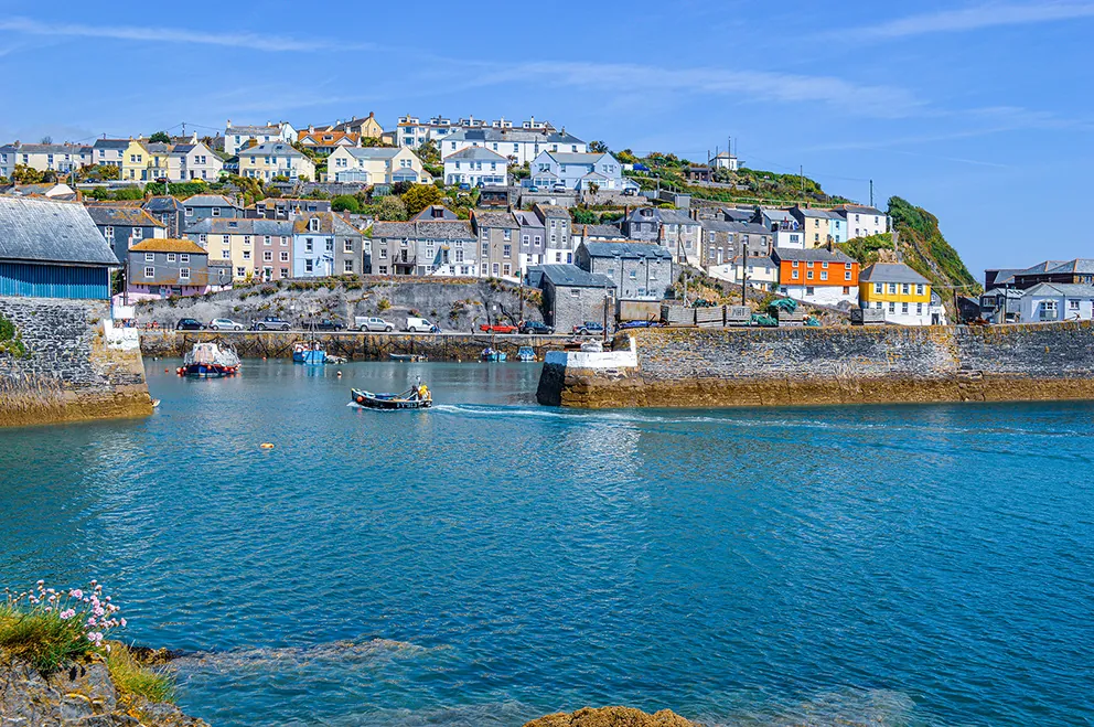 The image depicts a picturesque coastal town with colorful houses built on a hillside overlooking a harbor. The harbor features calm, clear blue water with a few small boats. The town appears to be a charming, quaint location, likely a popular spot for tourists and locals alike.