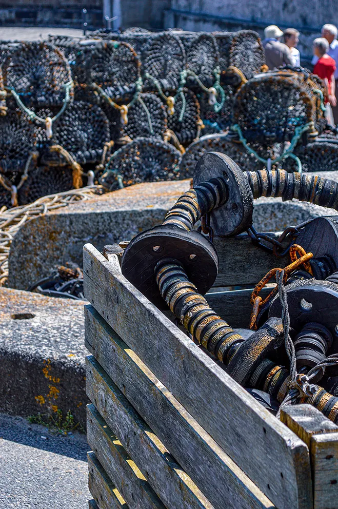 The image depicts a close-up view of a winch mechanism used in a fishing or maritime setting. The winch is attached to a weathered wooden platform, with ropes and chains coiled around it. In the background, there are stacks of lobster pots or similar fishing equipment, and a few people are visible, likely engaged in fishing-related activities.