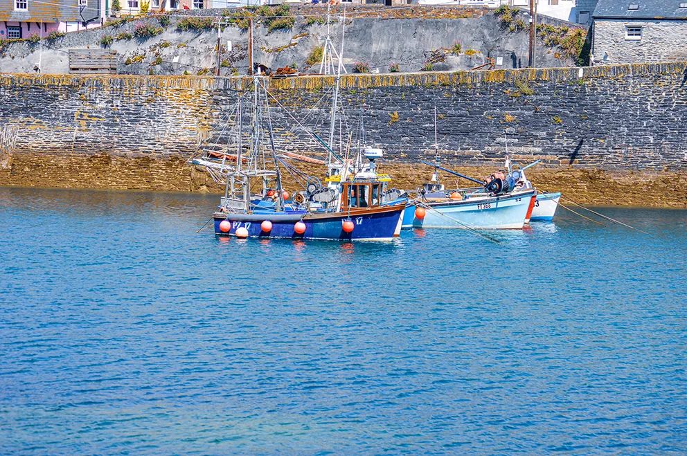 The image shows two small boats floating on calm blue water near a stone harbor wall. The boats are moored with ropes tied to buoys and appear to be fishing boats, as they have various fishing equipment on board. The harbor wall is made of large stones and has some vegetation growing on top. The scene suggests a peaceful, coastal setting, likely in a small harbor or fishing village.