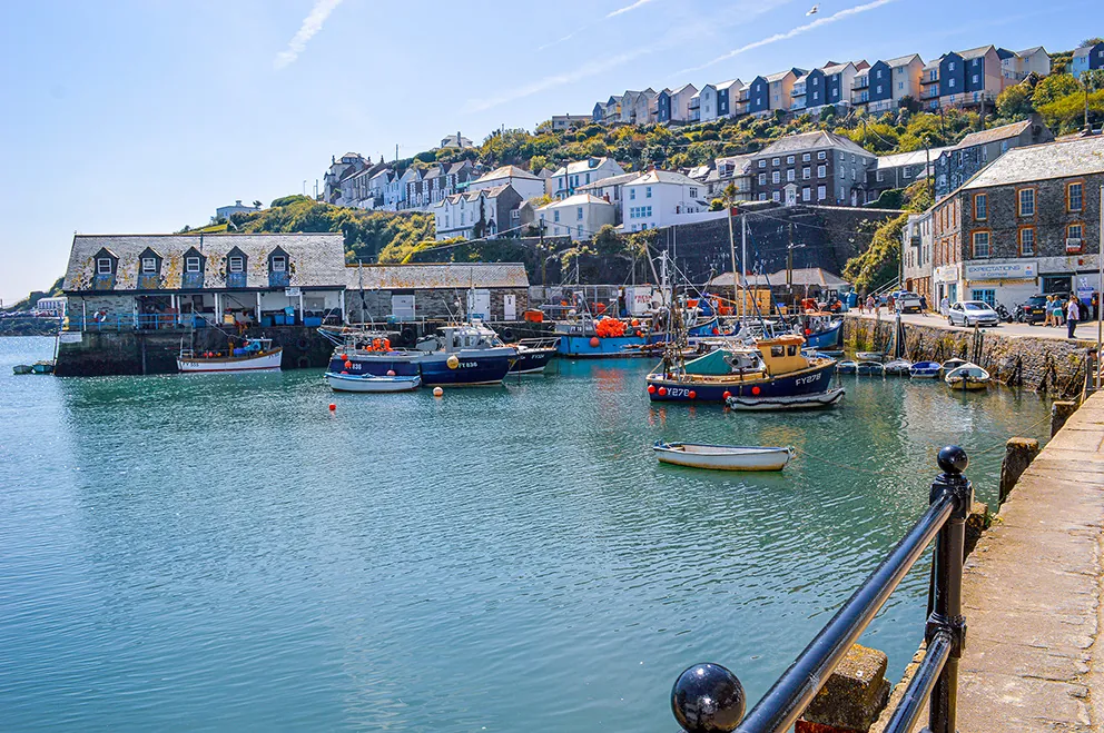 The image depicts a picturesque harbor scene with several boats docked in calm, clear water. The harbor is lined with charming buildings and houses that are built on a hillside, creating a colorful and quaint backdrop. The area appears to be a small, serene coastal village, likely a popular spot for fishing or tourism. The weather is clear, and the overall atmosphere is peaceful and inviting.