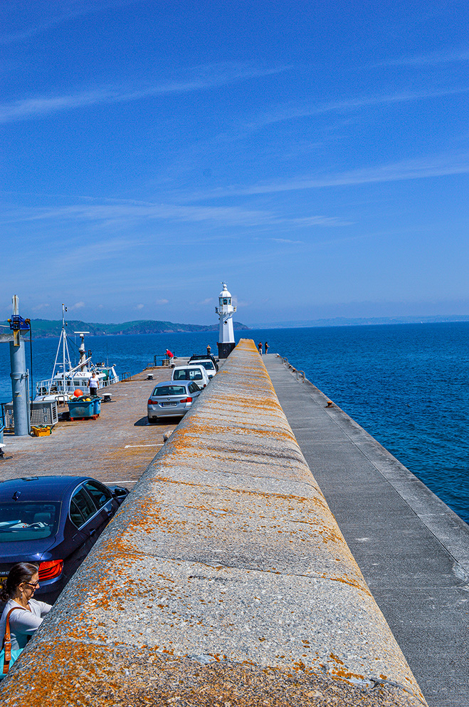 The image depicts a scenic view of a pier extending into a body of water, with a lighthouse at the end. Cars are parked along the pier, and a few people are visible enjoying the view. The sky is clear with a few clouds, and the water is calm, creating a serene atmosphere.