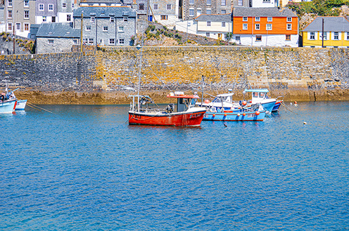 The image depicts a serene coastal scene with several boats anchored in a calm, blue harbor. The boats are moored close to a stone wall that lines the shore, behind which are colorful houses and buildings. The overall atmosphere is peaceful and picturesque, typical of a small coastal village.