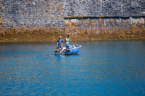 The image shows two people in a small boat on a body of water near a stone wall. They appear to be engaged in some activity, possibly related to fishing or maintenance, as one person is holding a tool or object.