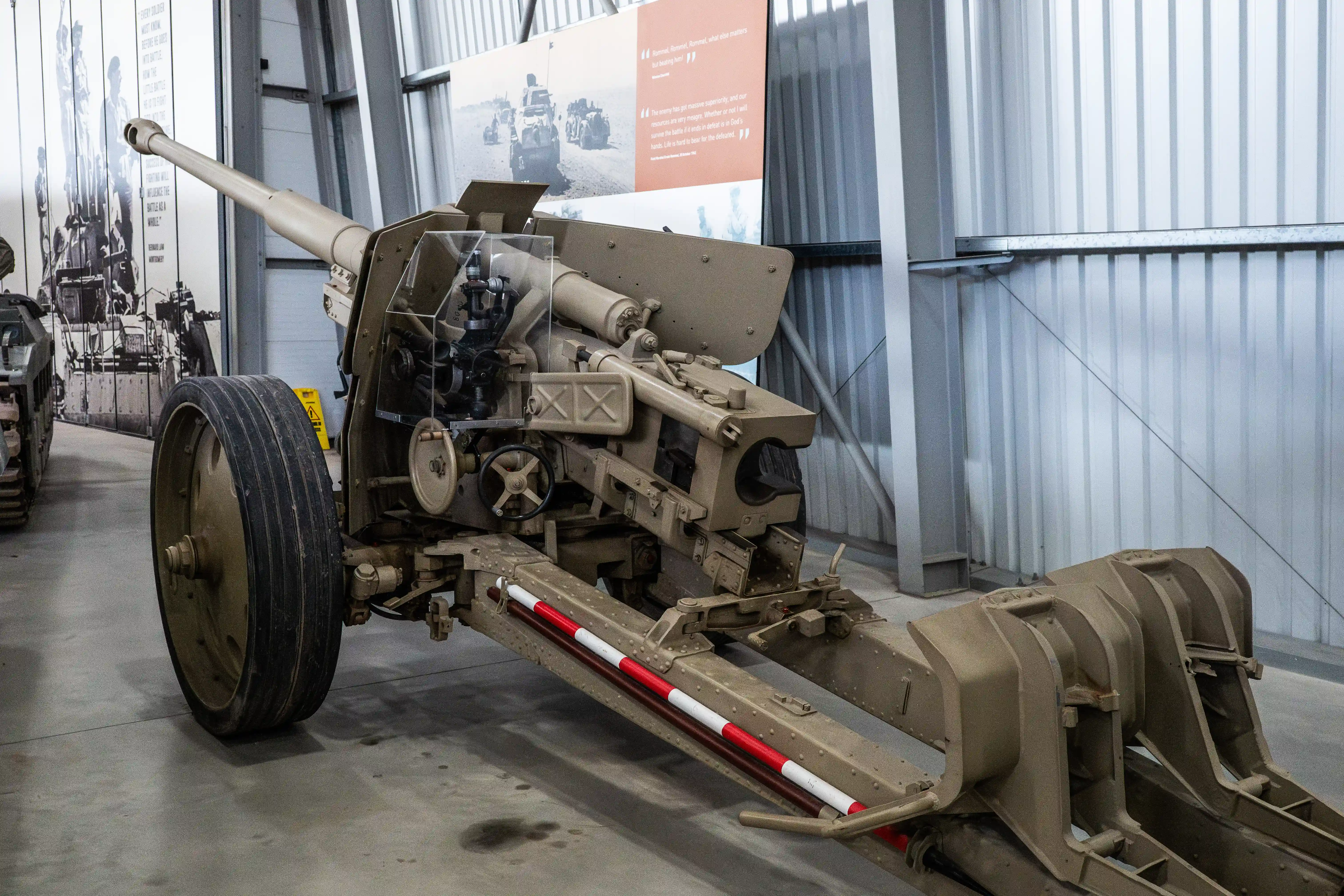 The image depicts a historical artillery piece displayed in a museum setting. The artillery piece is mounted on wheels and features a large barrel pointed upwards. The exhibit is housed in a modern facility with large windows and informative displays on the walls. The surroundings include other military equipment and informational posters, suggesting an educational focus on military history.