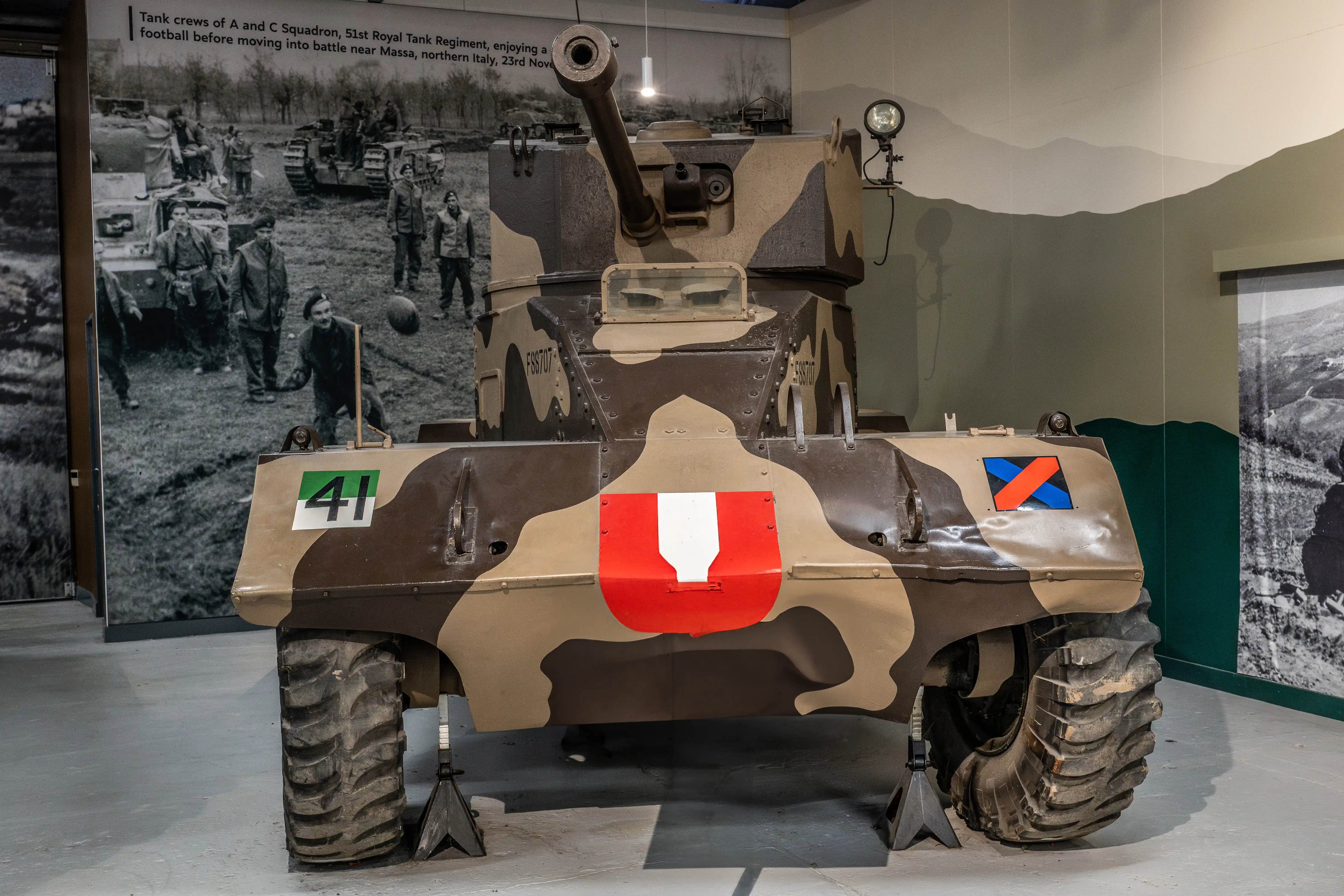 The image shows a military vehicle, specifically a tank, on display in a museum. The tank is painted with camouflage and has various markings, including a large red and white emblem on the front and smaller symbols on the sides. Behind the tank, there is a black-and-white photograph depicting tank crews from the 51st Royal Tank Regiment enjoying a game of football before heading into battle near Massa, northern Italy, on November 23rd.