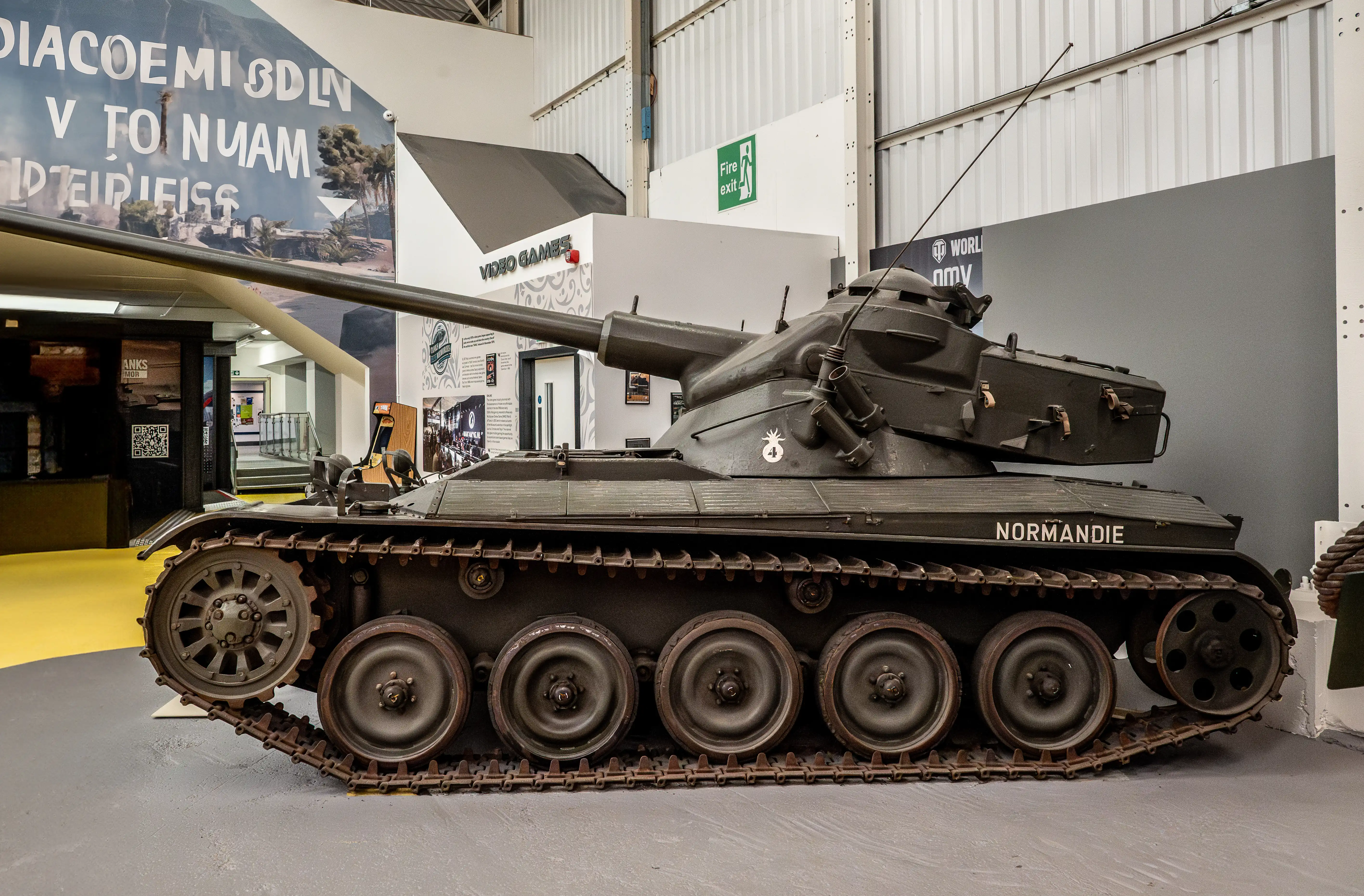 The image depicts a military tank named 'NORMANDIE' displayed indoors, likely in a museum or exhibition setting. The tank is positioned on a flat surface with tracks visible, and it features a large gun barrel extending from its turret. Surrounding the tank are various informational displays and signs, including a fire exit sign and a QR code on a nearby wall. The environment suggests an educational exhibit focused on military history, specifically related to World War II.