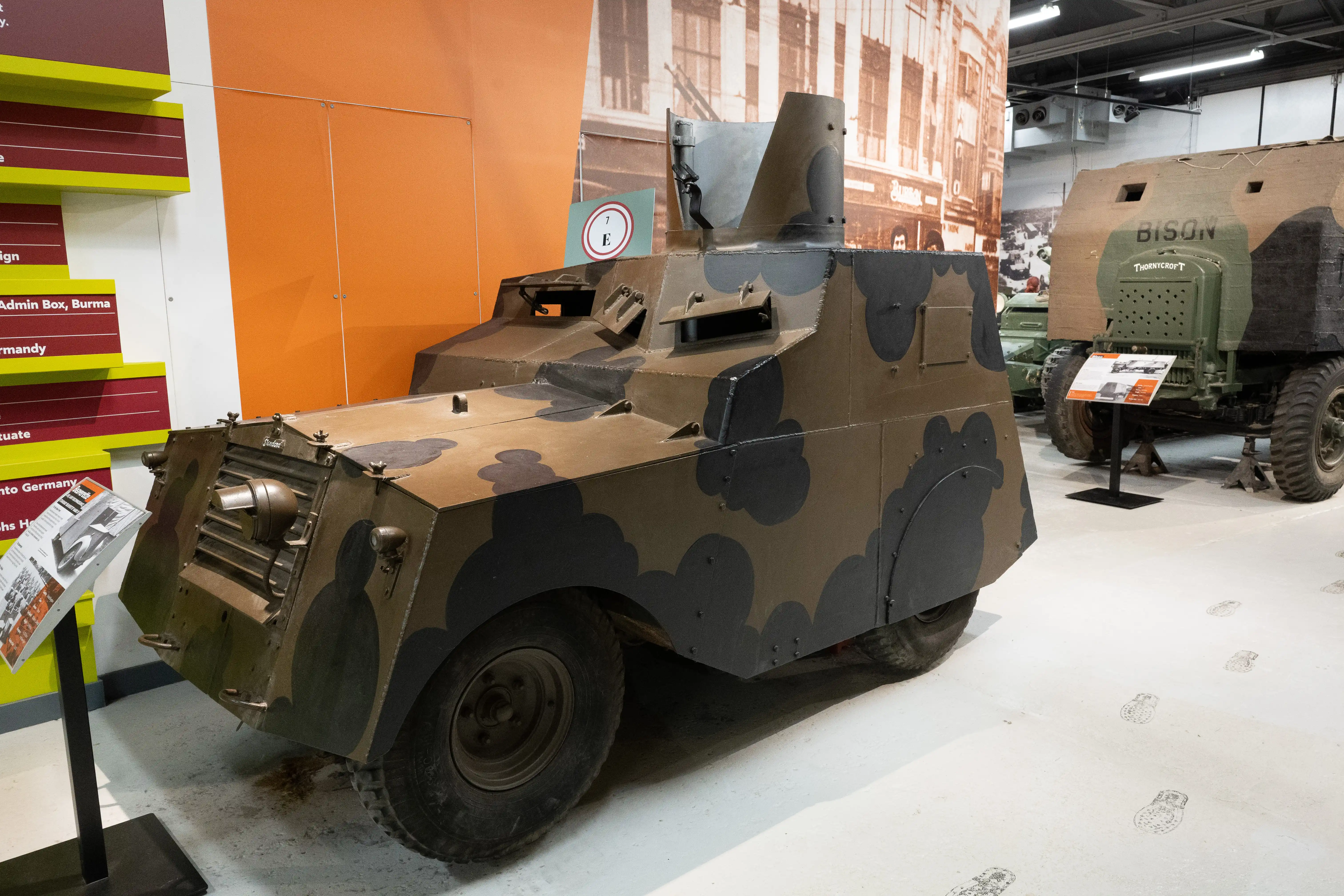 The image depicts a military exhibition featuring two vintage armored vehicles. The foreground vehicle is a small, camouflaged armored car with a turret, likely used in reconnaissance or light combat roles. The background vehicle is a larger, green armored truck labeled 'Bison' with the manufacturer's name 'Thornycroft' visible. Both vehicles are displayed indoors, with informational placards in front of them, providing context and historical details about their use and significance.