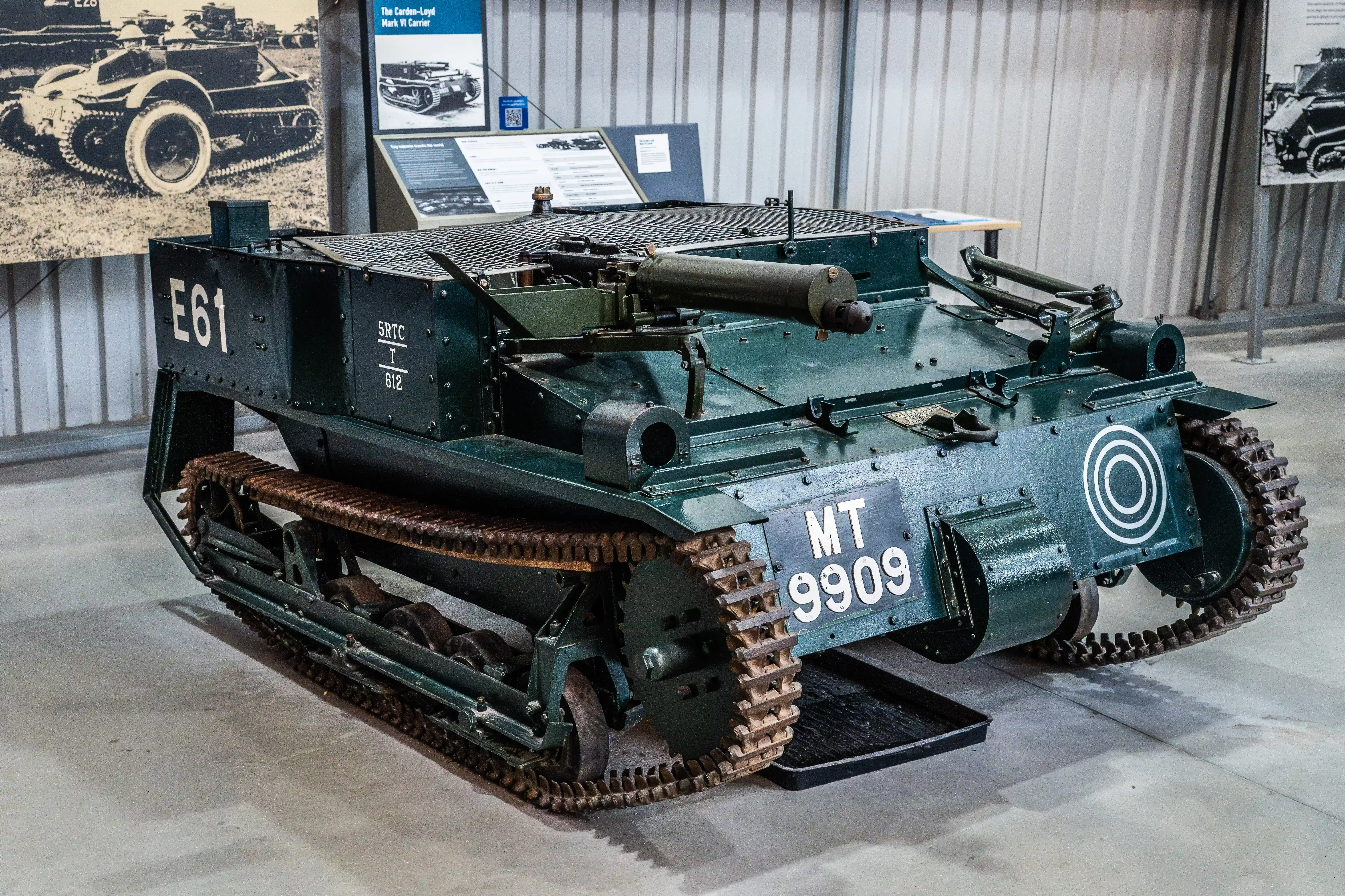 The image depicts a historical military tracked vehicle, specifically a Carden-Loyd Mark VI Tankette, on display in a museum setting. The vehicle is painted in a dark green color and has the markings 'E61' and 'MT 9909' on its side. The tankette features a mesh-covered rear compartment and a cylindrical object on top, likely a mounting for a machine gun. The background includes informational displays and black-and-white photographs of similar vehicles, providing context and historical information about the vehicle's design and usage.