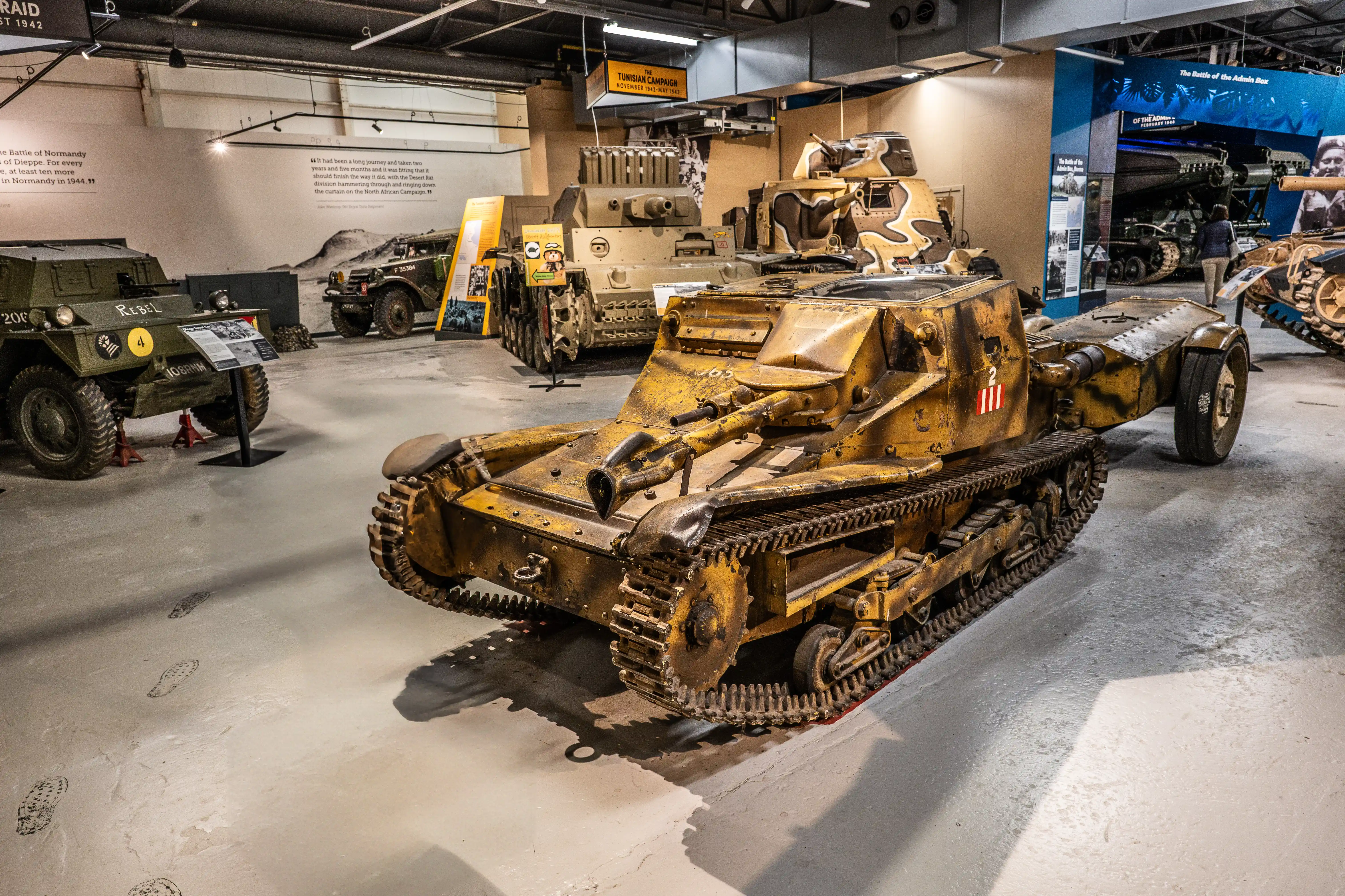 The image depicts a military museum exhibit featuring various armored vehicles, including tanks and reconnaissance vehicles, from different historical periods. The vehicles are displayed in a well-lit indoor space with informational placards and background displays providing context about specific battles and campaigns, such as the Battle of Normandy and the Tunisian Campaign.