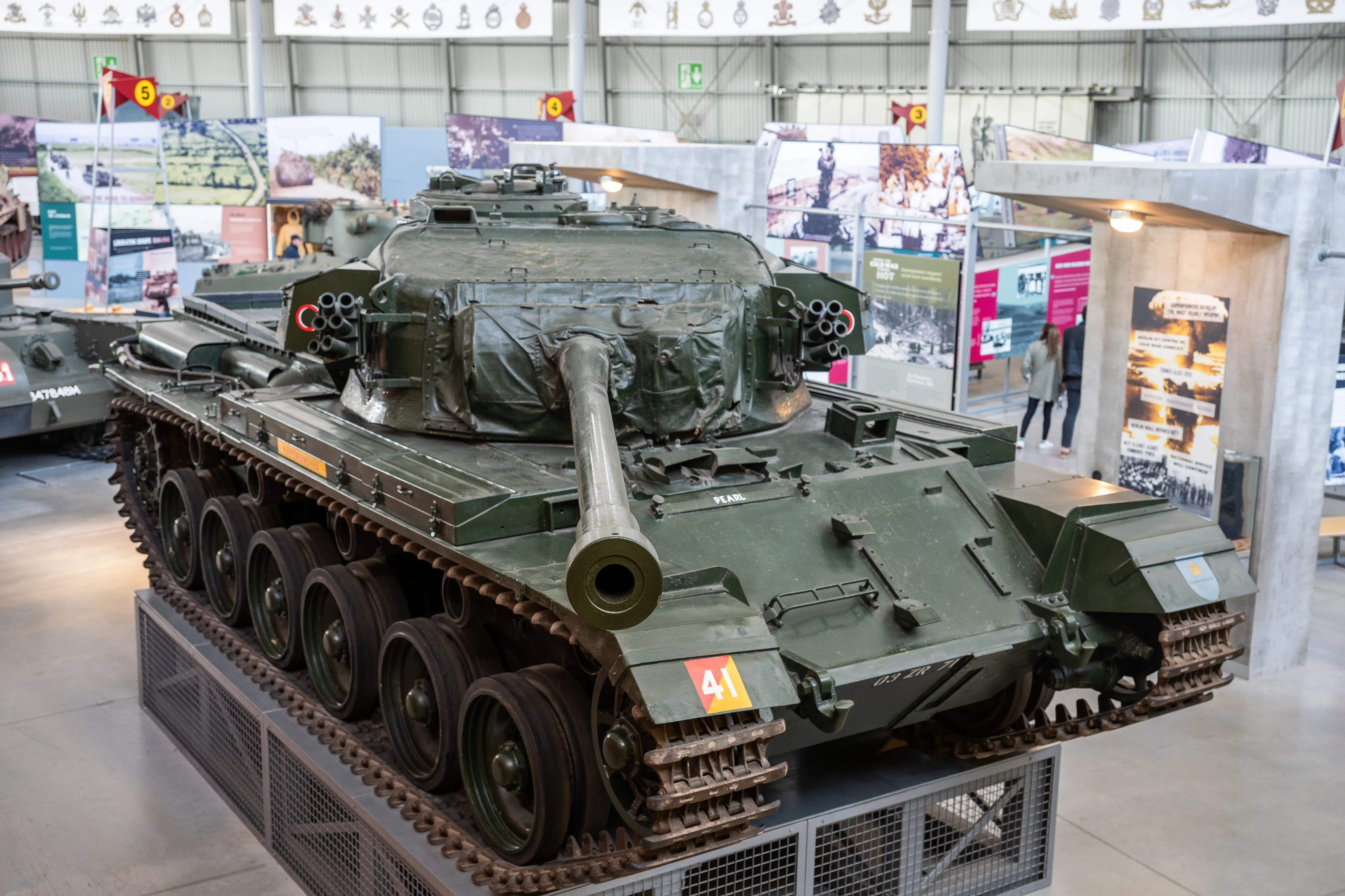 The image depicts a military tank displayed in a museum setting. The tank is painted in a green camouflage pattern and features a large cannon mounted on its turret. The surroundings include various informational displays and banners, suggesting an exhibition focused on military history or armored vehicles. The tank is positioned on a raised platform with protective barriers around it, indicating it is a significant exhibit. The overall scene suggests an educational environment aimed at showcasing historical military equipment.
