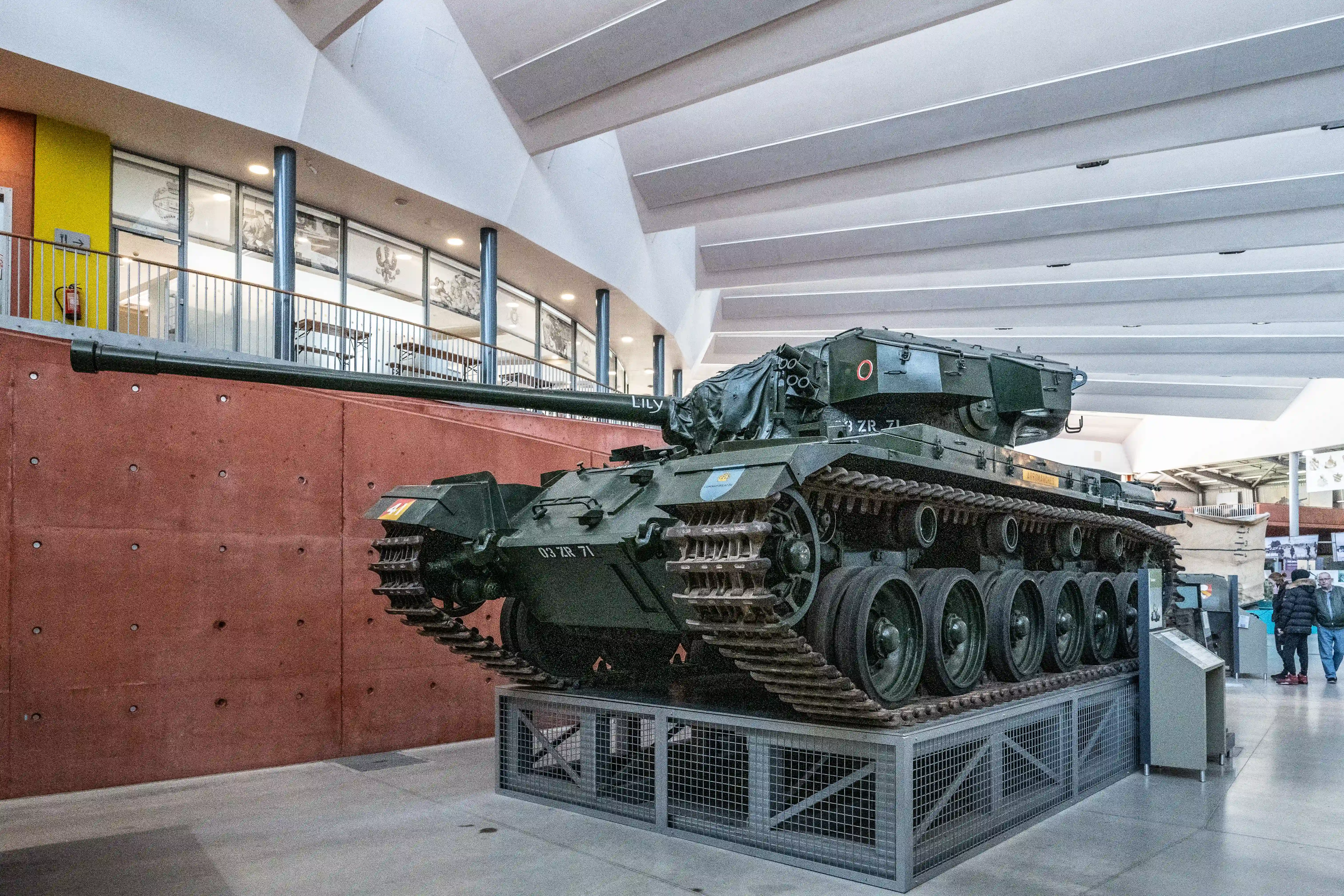 The image shows a military tank displayed indoors, likely in a museum or exhibition setting. The tank is mounted on a platform with a protective metal grate surrounding it. The interior space features modern architectural elements, including a high ceiling with linear lighting, large windows, and a balcony with railings. There are informational displays and a few people observing the exhibit.