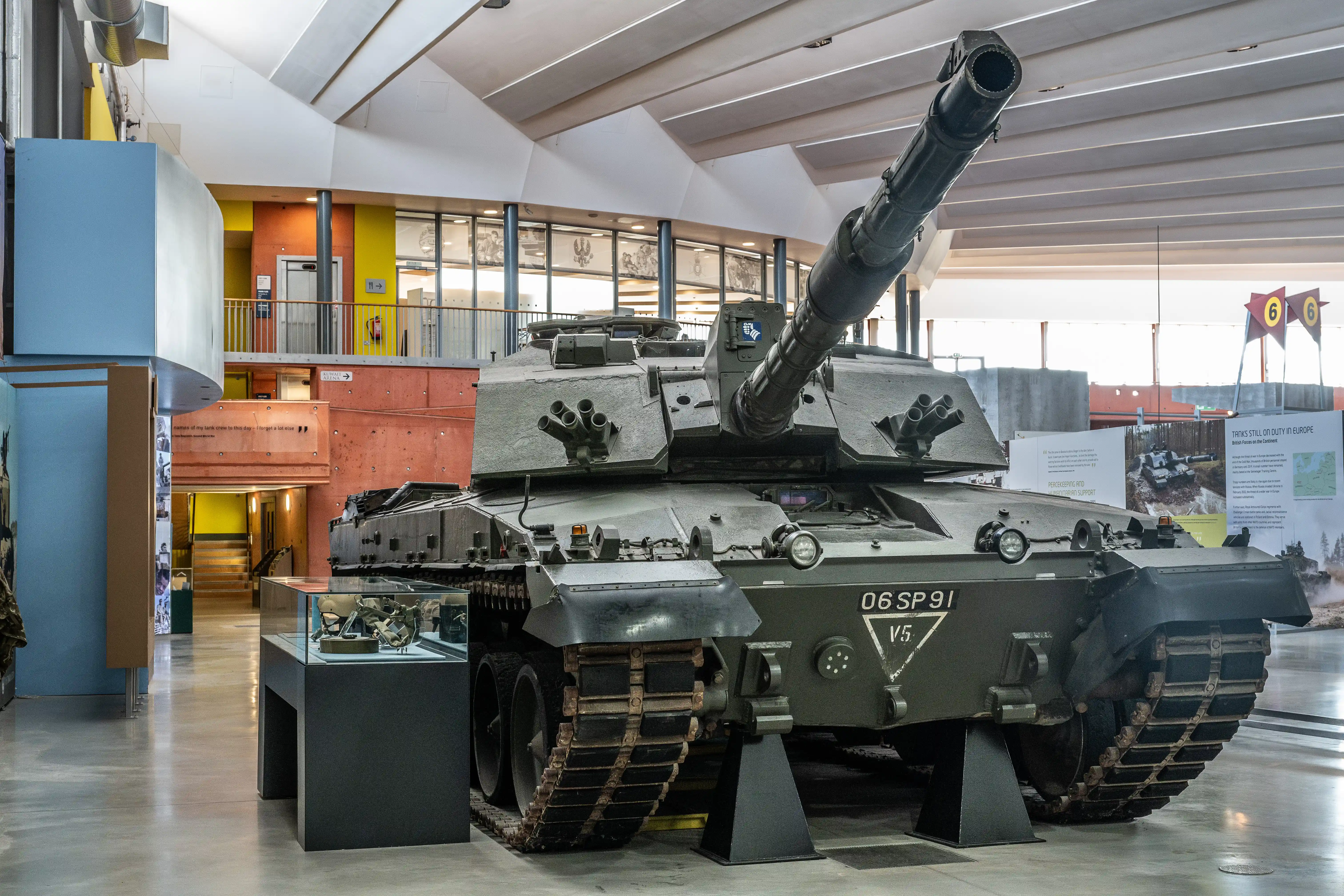 The image shows a military tank displayed in a museum setting. The tank is labeled '06 SP 91 V5' and is positioned indoors with various informational displays and artifacts around it. The museum has a modern design with large windows and multiple floors, providing a well-lit and spacious environment for visitors to explore.
