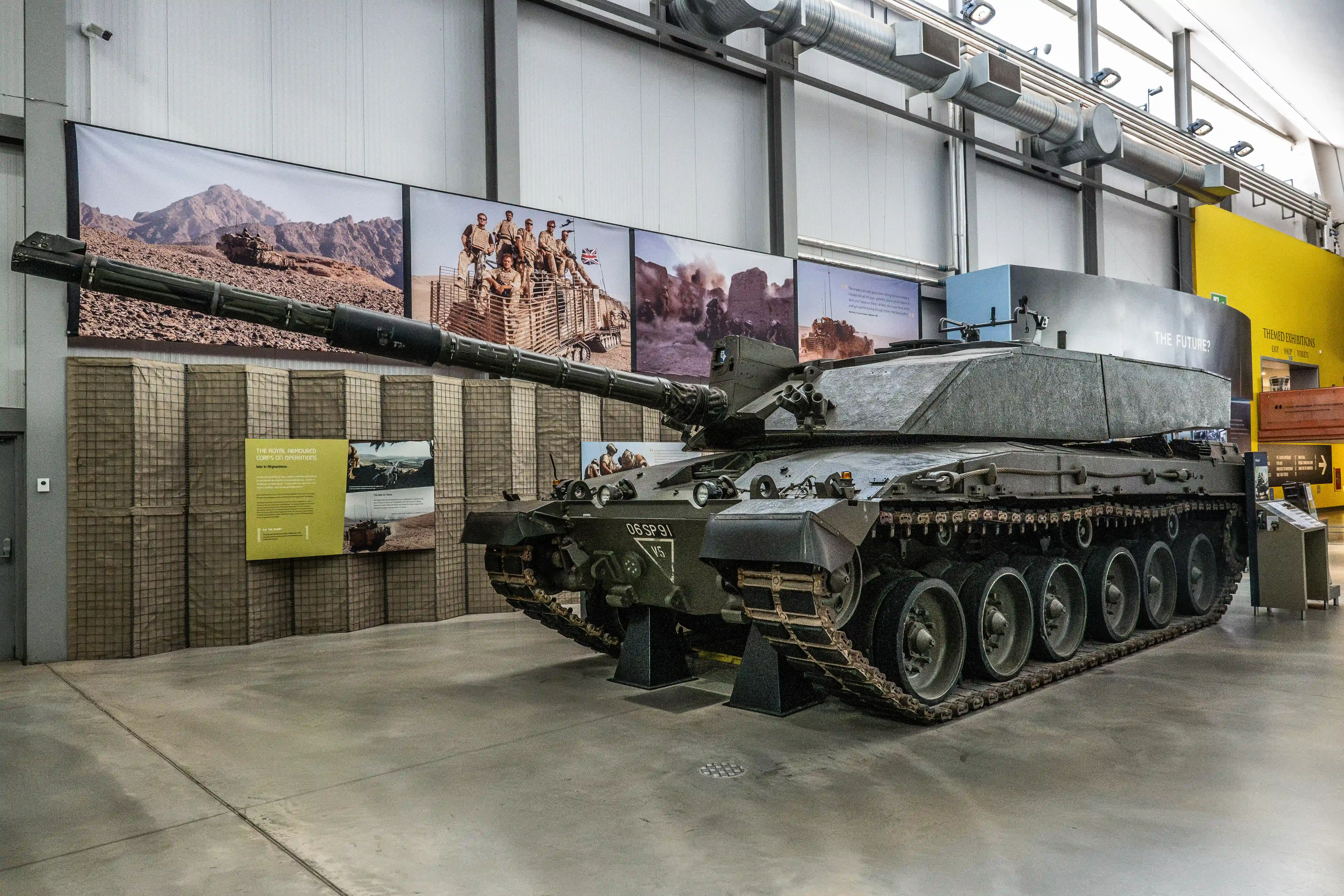 The image depicts a military tank displayed in a museum setting. The tank is prominently positioned in the center of the image, with its large gun barrel extending forward. The surroundings include informational panels and photographs related to the tank and its historical context, suggesting that this exhibit is part of a larger display about military history. The environment appears to be well-lit and organized, indicating a well-maintained exhibition space.