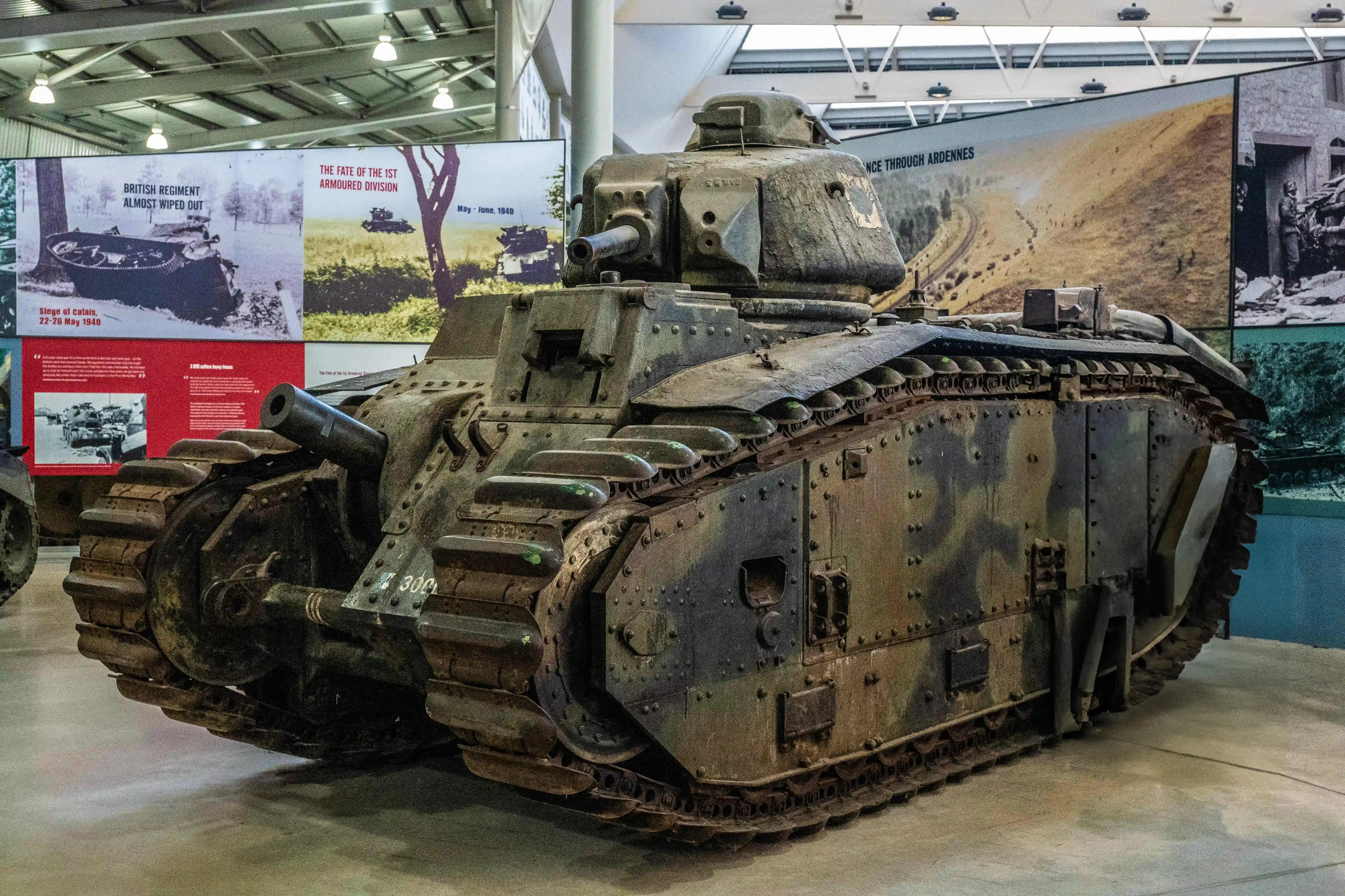 The image depicts a historical military tank displayed in a museum setting. The tank is an example of early 20th-century armored warfare technology. Surrounding informational panels provide context about significant battles and events involving the tank, such as the Siege of Calais in 1940 and the fate of the 1st Armoured Division. The display highlights the tank's role and the historical significance of the battles it participated in.