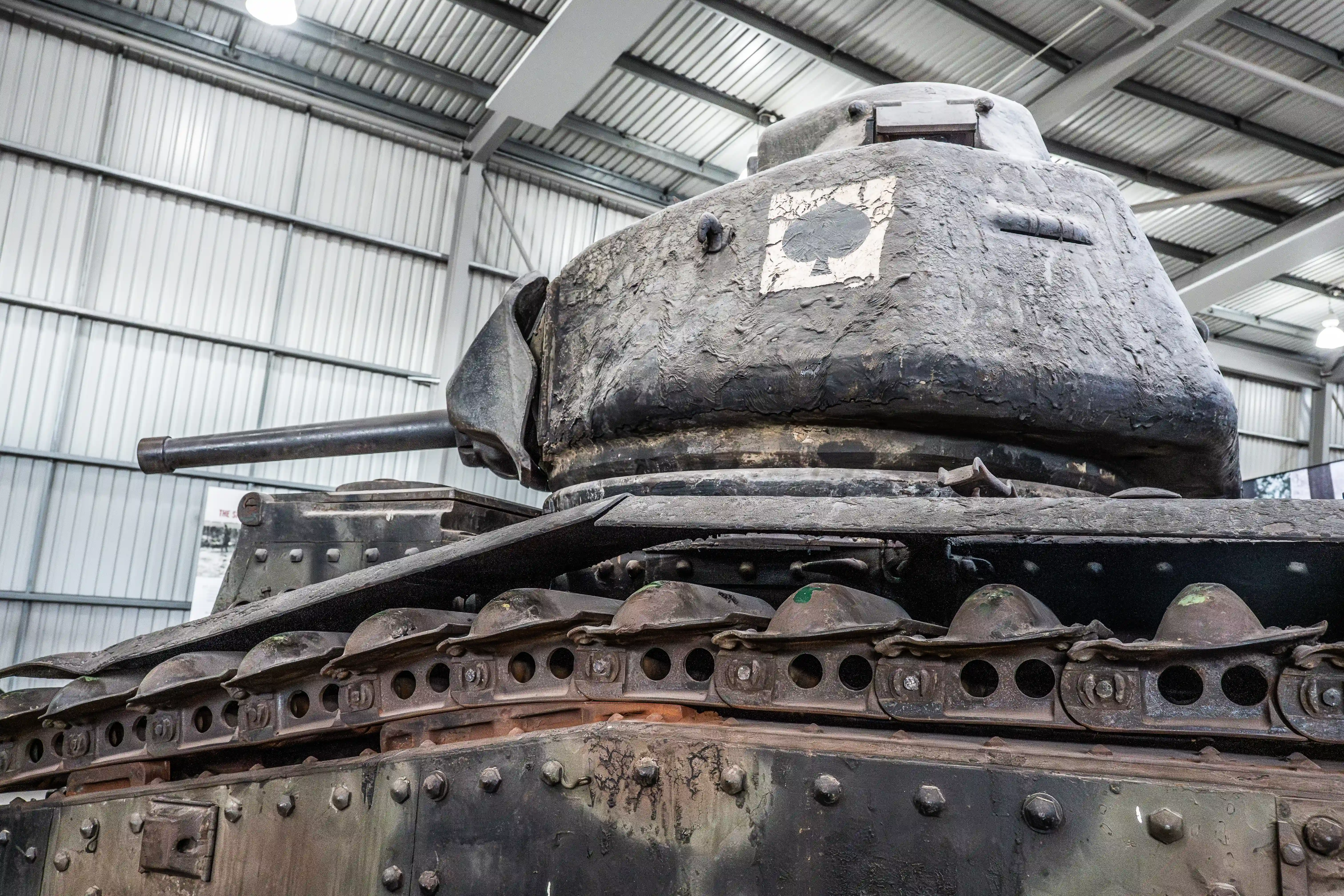 The image shows a close-up view of an old, weathered tank, likely from a historical period, housed in a large, modern warehouse or museum setting. The tank's turret and armor plating are prominently displayed, showing signs of wear and rust. The interior of the warehouse is visible in the background, with a high ceiling and metal siding