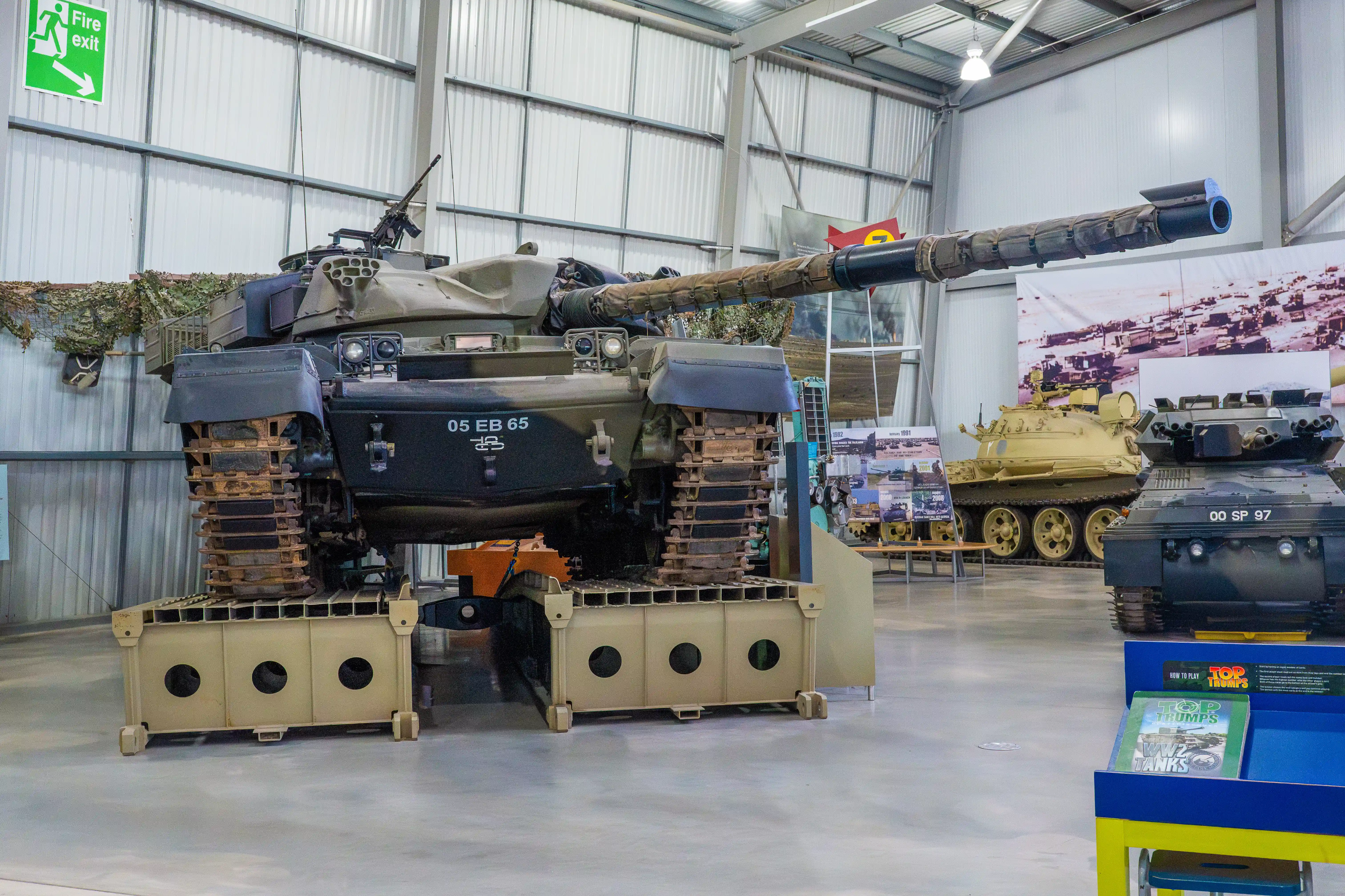 The image depicts an indoor exhibition of military tanks. The main tank in the foreground is elevated on supports, showcasing its underside. Other tanks are also on display in the background. The setting appears to be a well-lit, spacious hangar with informational displays and a fire exit sign visible. The exhibition likely focuses on military vehicles, providing educational insights into their design and historical significance.