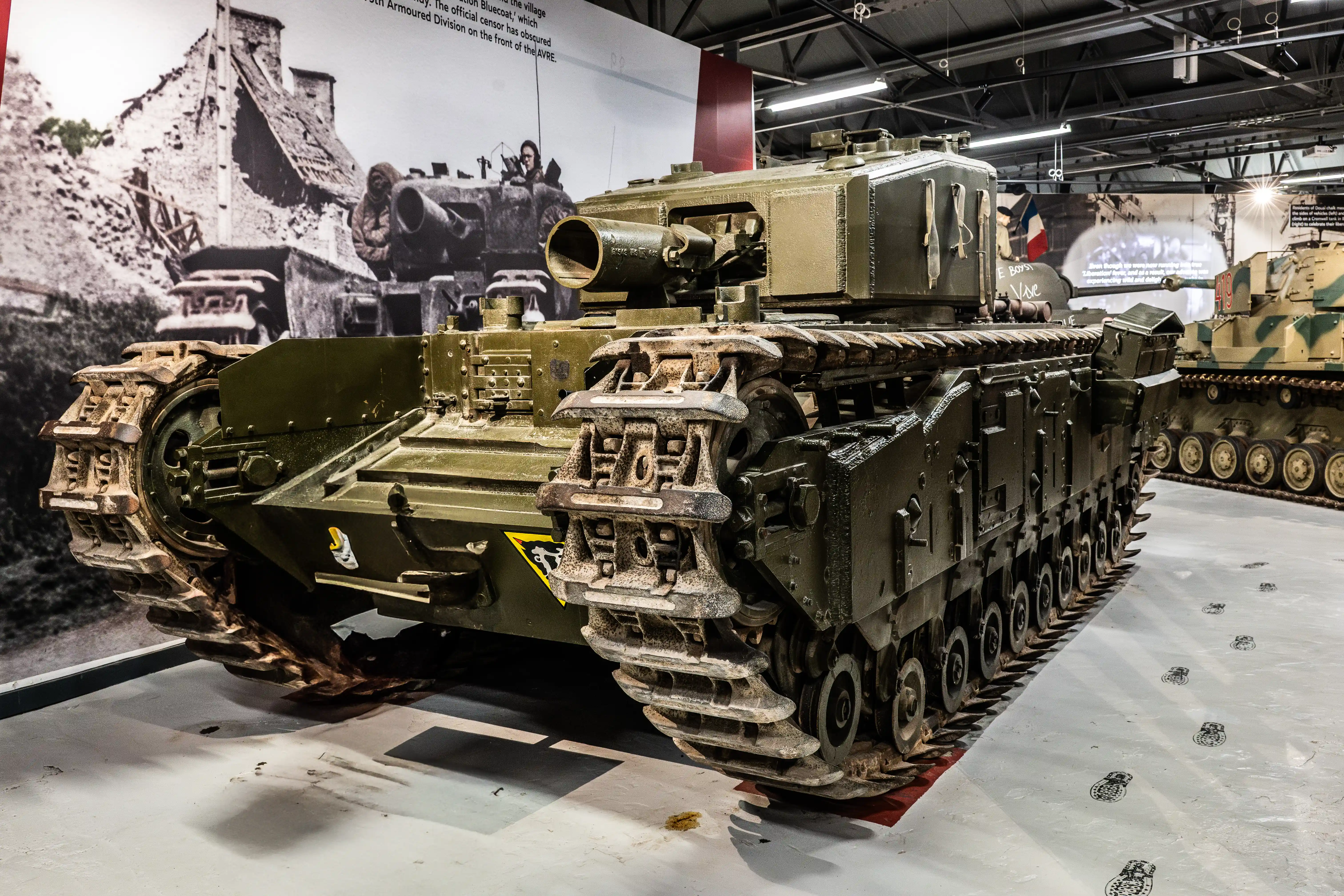 The image depicts a military tank on display at an exhibition, showcasing historical armored vehicles. The tank is prominently featured in the foreground, with another tank visible in the background. The display includes large black-and-white photographs of soldiers and military operations, providing a historical context. The setting appears to be indoors, with informative text panels explaining the significance of the vehicles and events depicted.