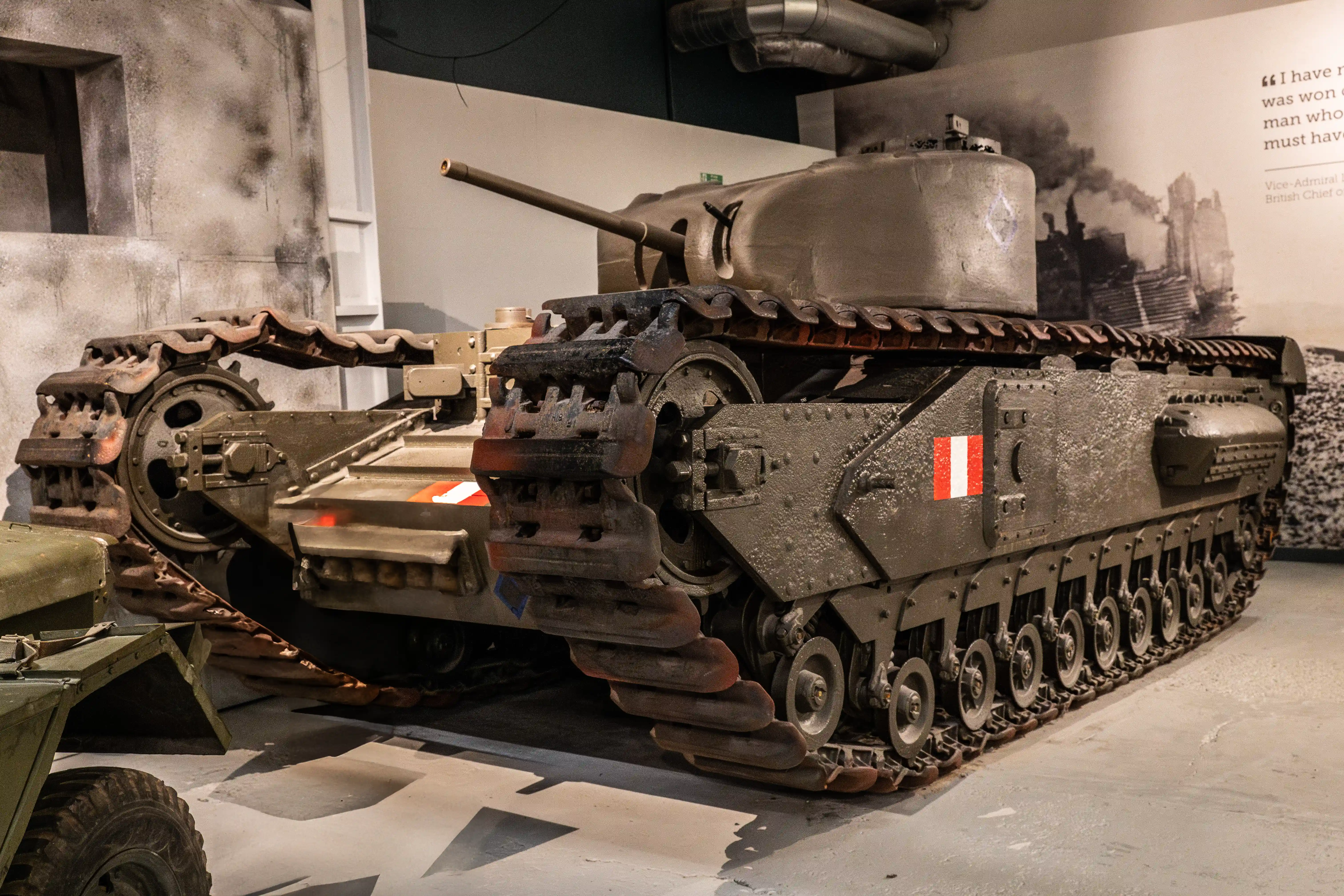 The image depicts a military tank on display at an exhibition, showcasing historical armored vehicles. The tank is prominently featured in the foreground, with another tank visible in the background. The display includes large black-and-white photographs of soldiers and military operations, providing a historical context. The setting appears to be indoors, with informative text panels explaining the significance of the vehicles and events depicted.