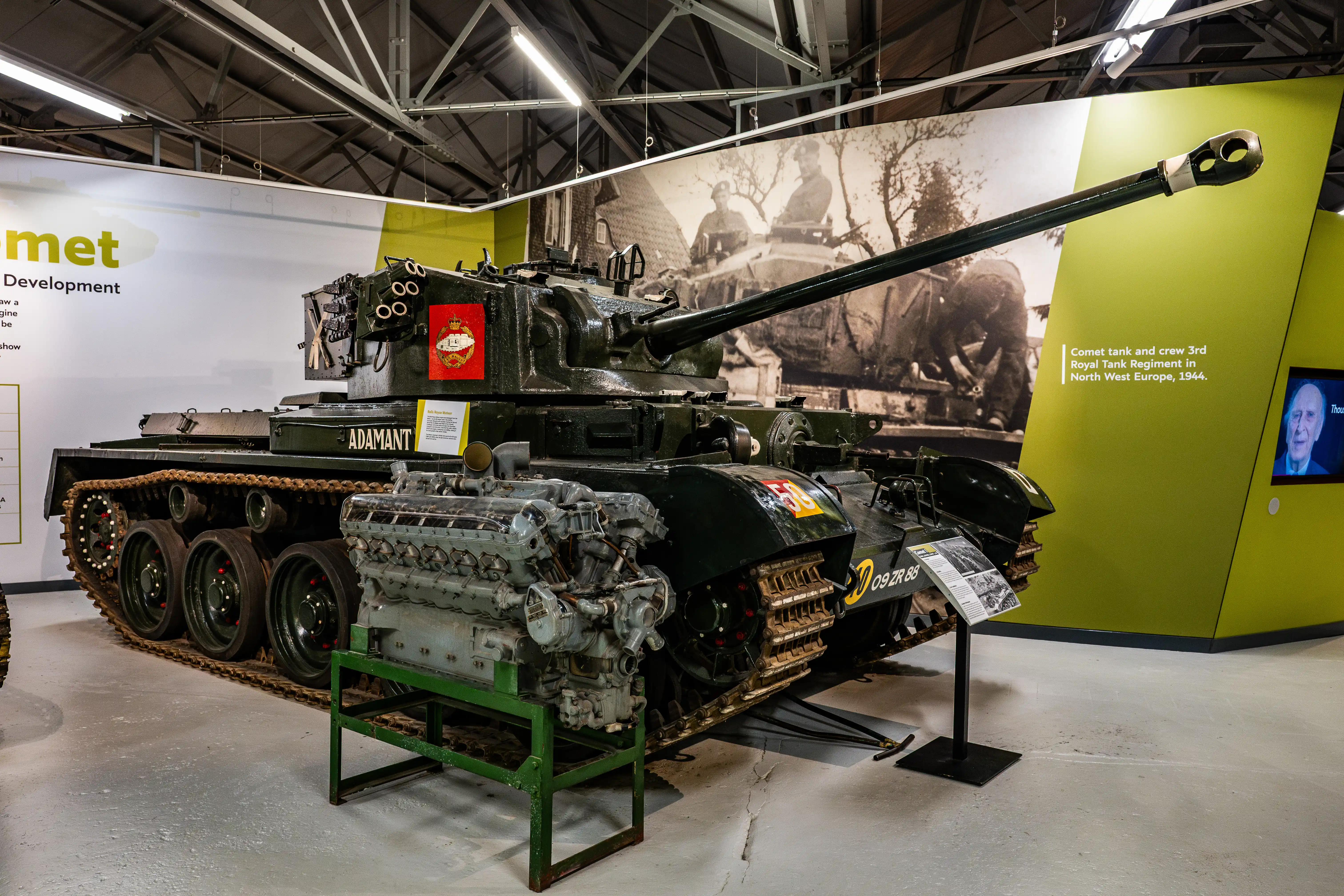 The image depicts a Comet tank and its crew from the 3rd Royal Tank Regiment in North West Europe, 1944. The tank is displayed in a museum setting, showcasing its development and historical significance. The exhibit includes the tank's engine and detailed information about its role and specifications.