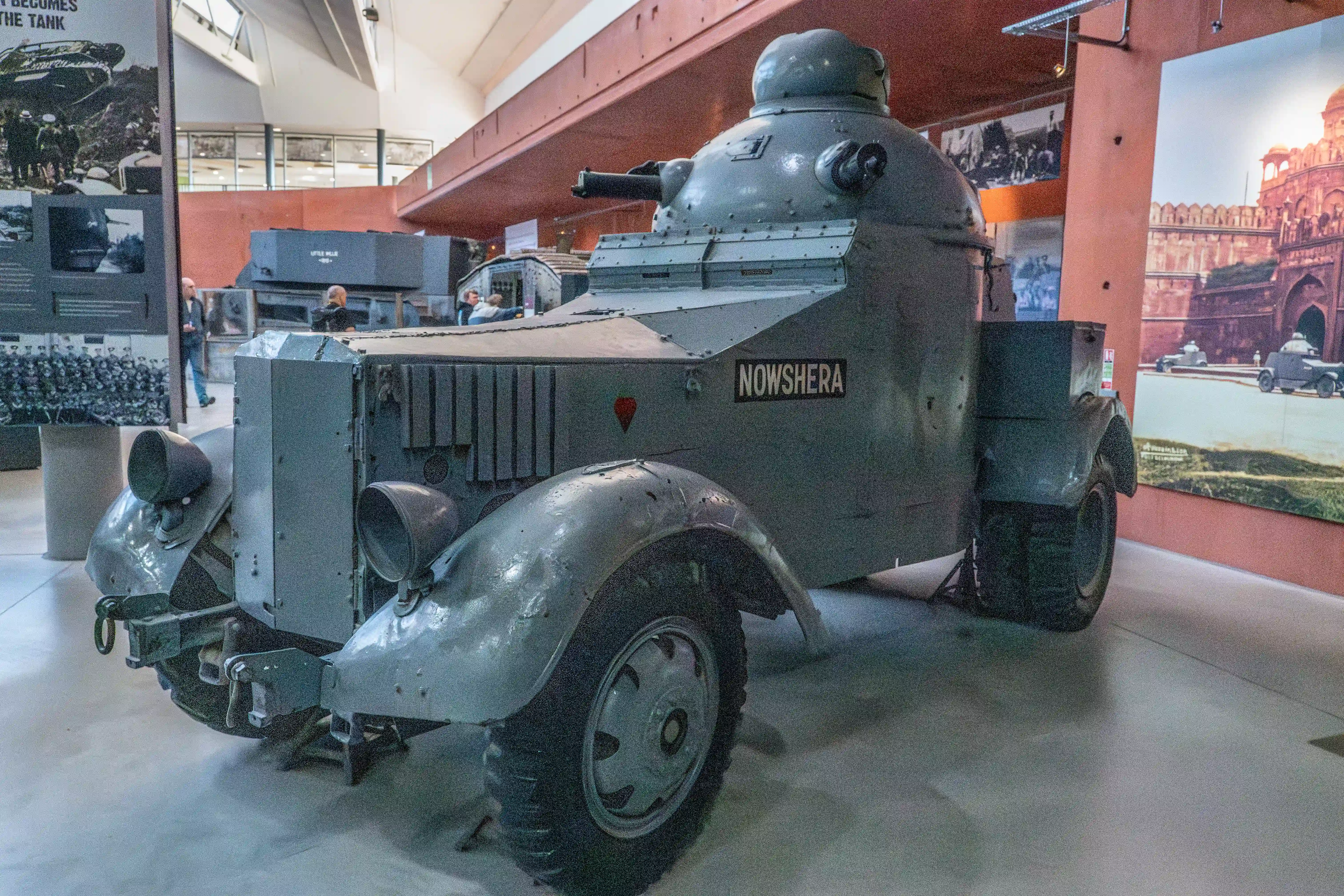 The image depicts an armored vehicle named 'Nowshera' on display in a museum. The vehicle has a rugged, metallic exterior with a turret on top, equipped with a cannon. It is displayed in an indoor exhibit area with historical photographs and informational panels in the background, suggesting its historical significance and use in military contexts.