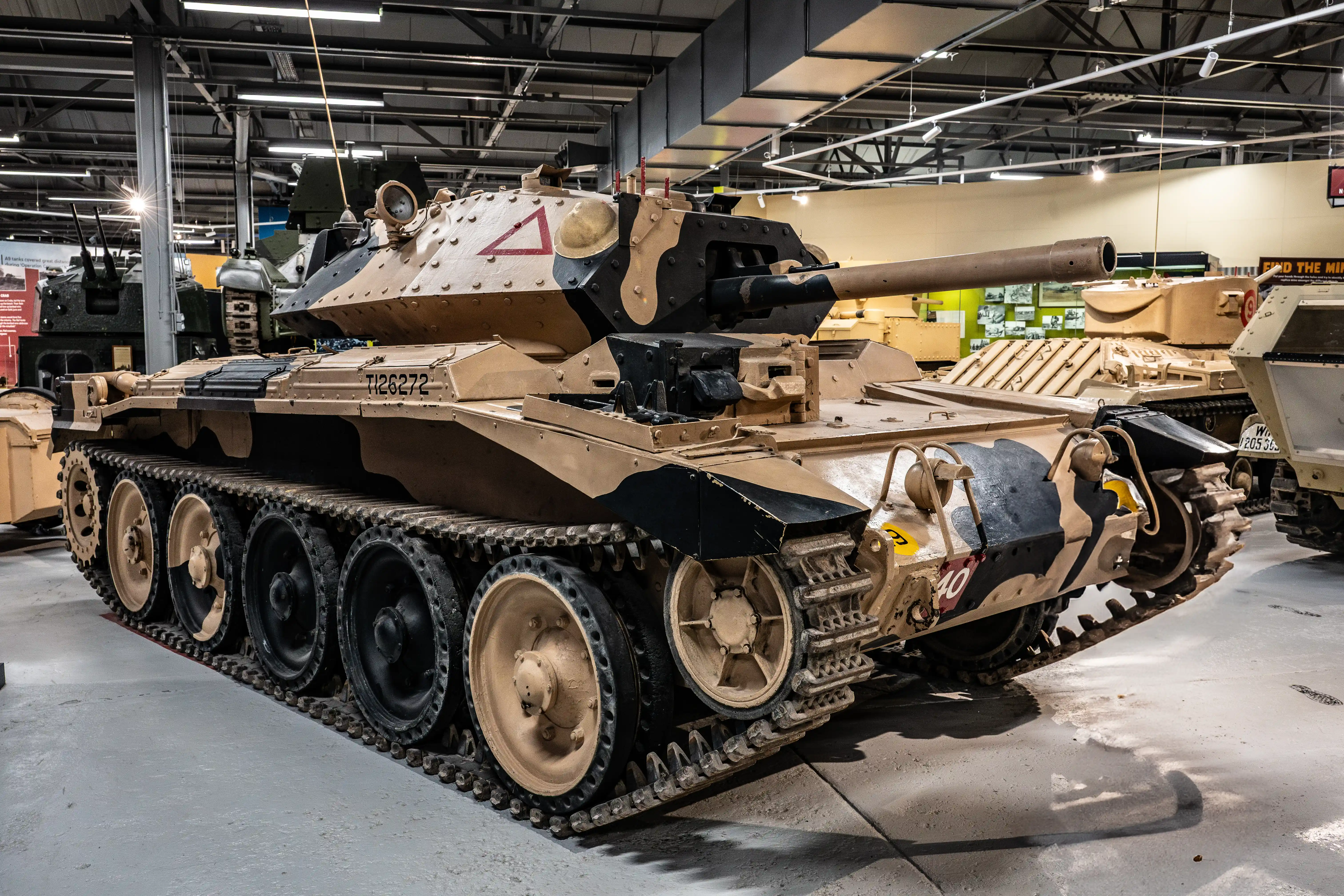 The image depicts a tank, specifically a tank museum exhibit. The tank is a World War II-era tank, likely a British Comet tank, identifiable by its distinctive shape and markings. The tank is displayed indoors, with various other military vehicles visible in the background. The tank has a camouflage paint scheme and is equipped with a large main gun. The setting appears to be a well-lit, spacious museum with informational displays and signage.