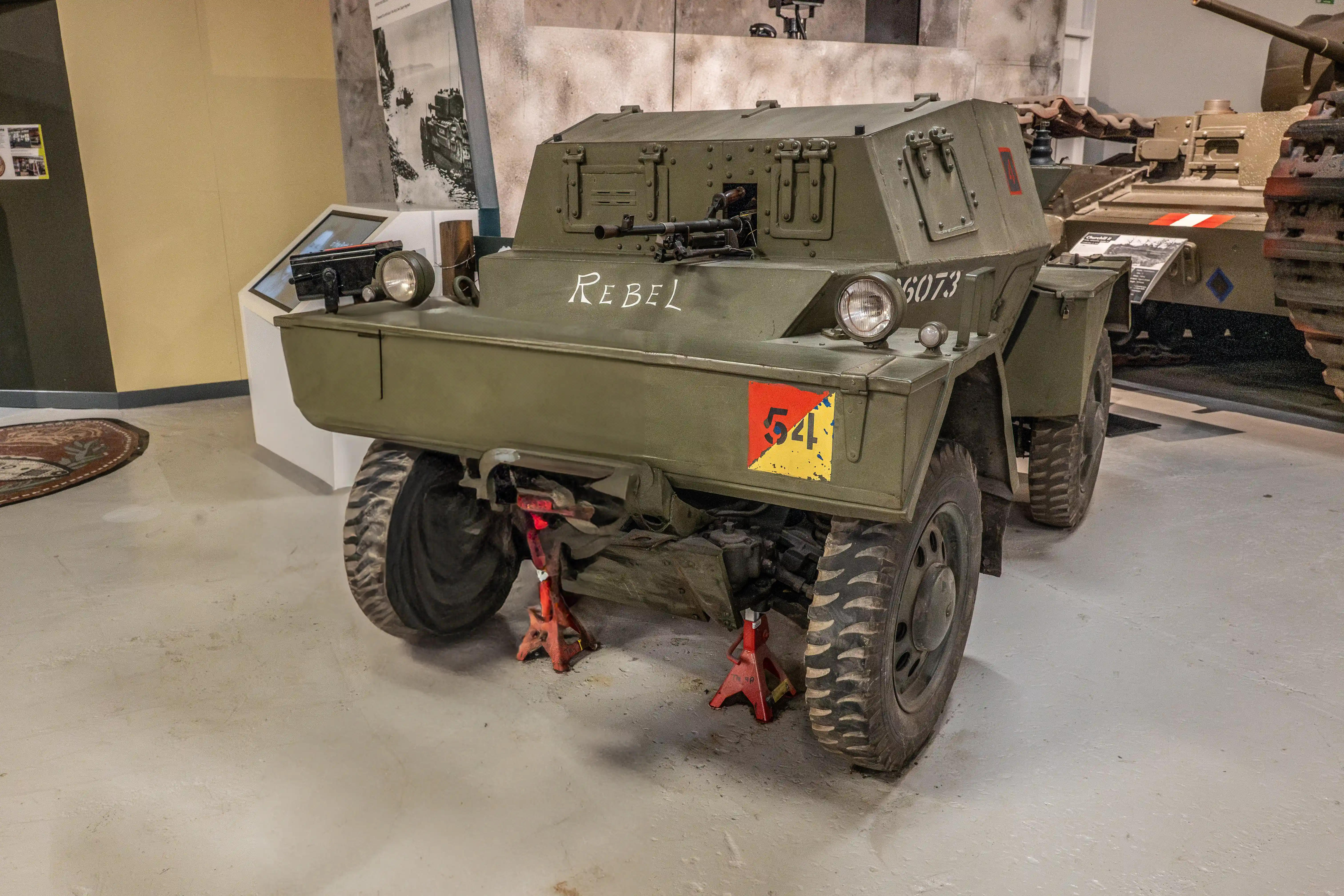 The image depicts a military vehicle, specifically an armored car, on display in a museum or exhibition setting. The vehicle is painted in a military green color and has various markings, including the word 'Rebel' and the number '6073' on its front. It is elevated on red jack stands, suggesting it is being maintained or displayed in a stationary position. The vehicle is equipped with heavy-duty tires and has a machine gun mounted on top. The background includes other military artifacts and displays, indicating a historical or military museum context.
