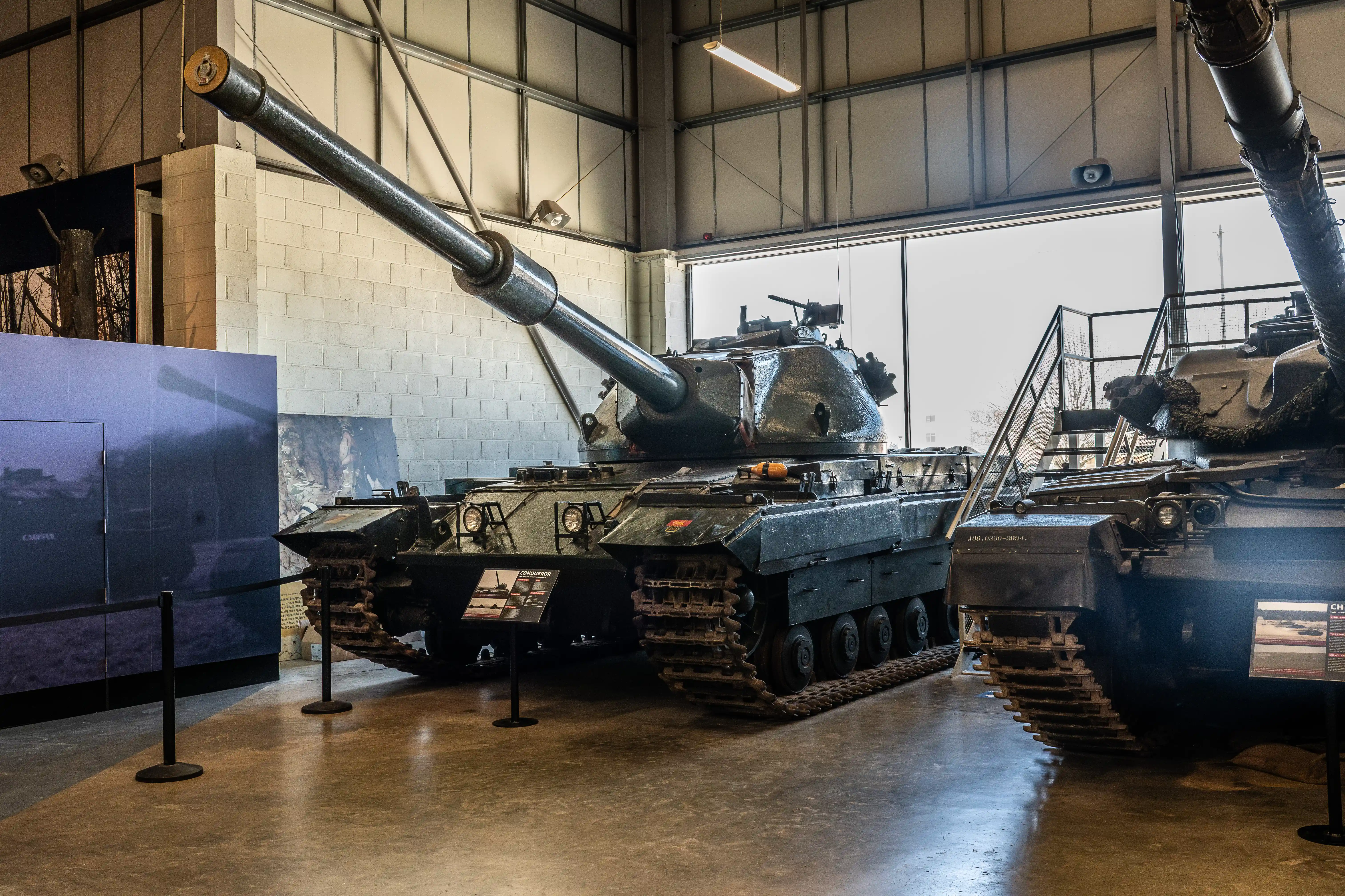 The image depicts a military museum exhibit featuring several tanks. The central tank has a large, prominent gun barrel, and there are informational placards in front of each tank, likely providing details about their history and specifications. The exhibit is housed in a spacious, well-lit hangar with large windows, allowing natural light to illuminate the display area.