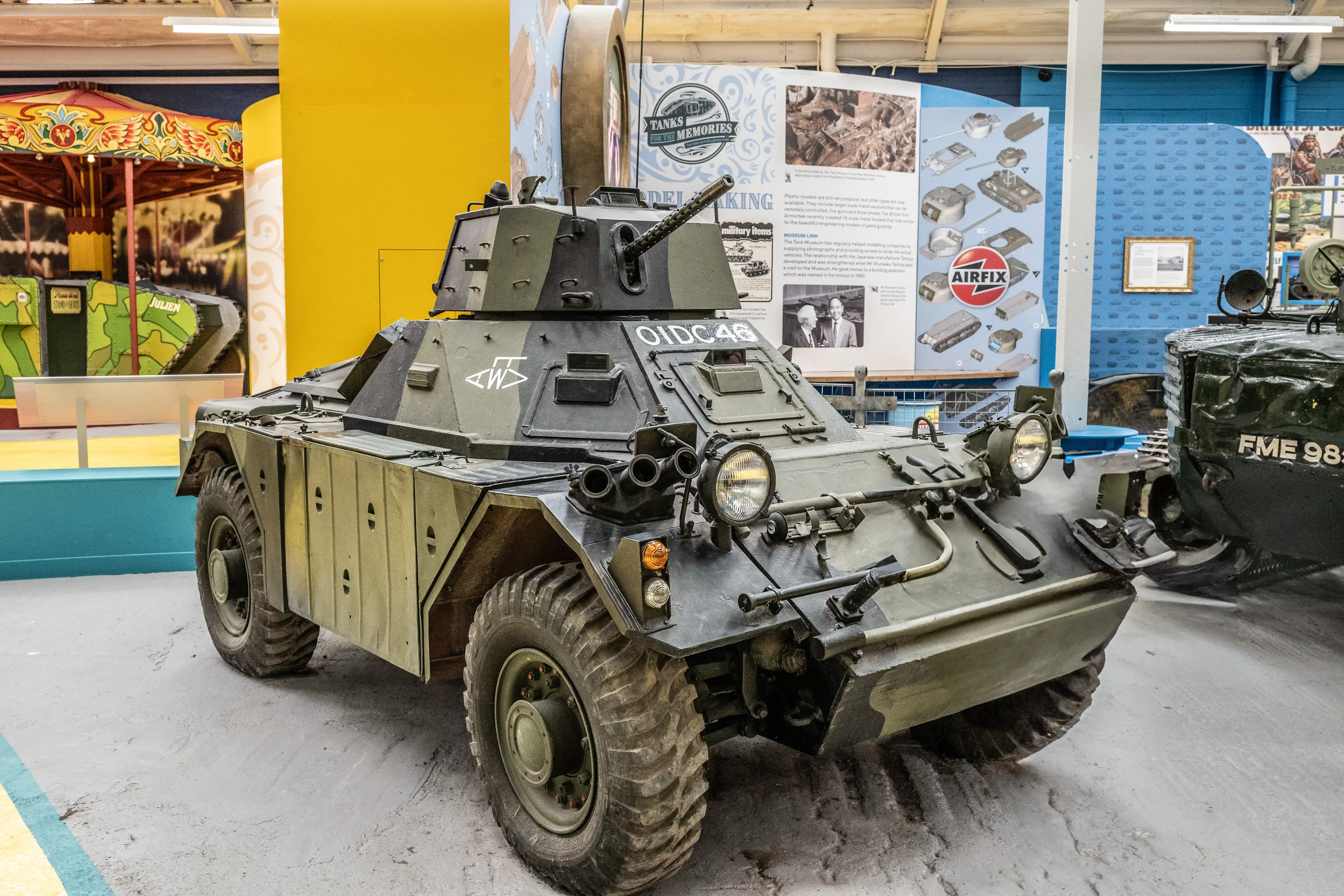 The image depicts a military armored vehicle on display in a museum. The vehicle, labeled 'OILCAR,' is an armored car with various military features such as mounted lights, a machine gun, and reinforced plating. The display area includes informational posters about the vehicle and related exhibits. The setting appears to be part of a larger exhibit on military history, with other vehicles and displays visible in the background.