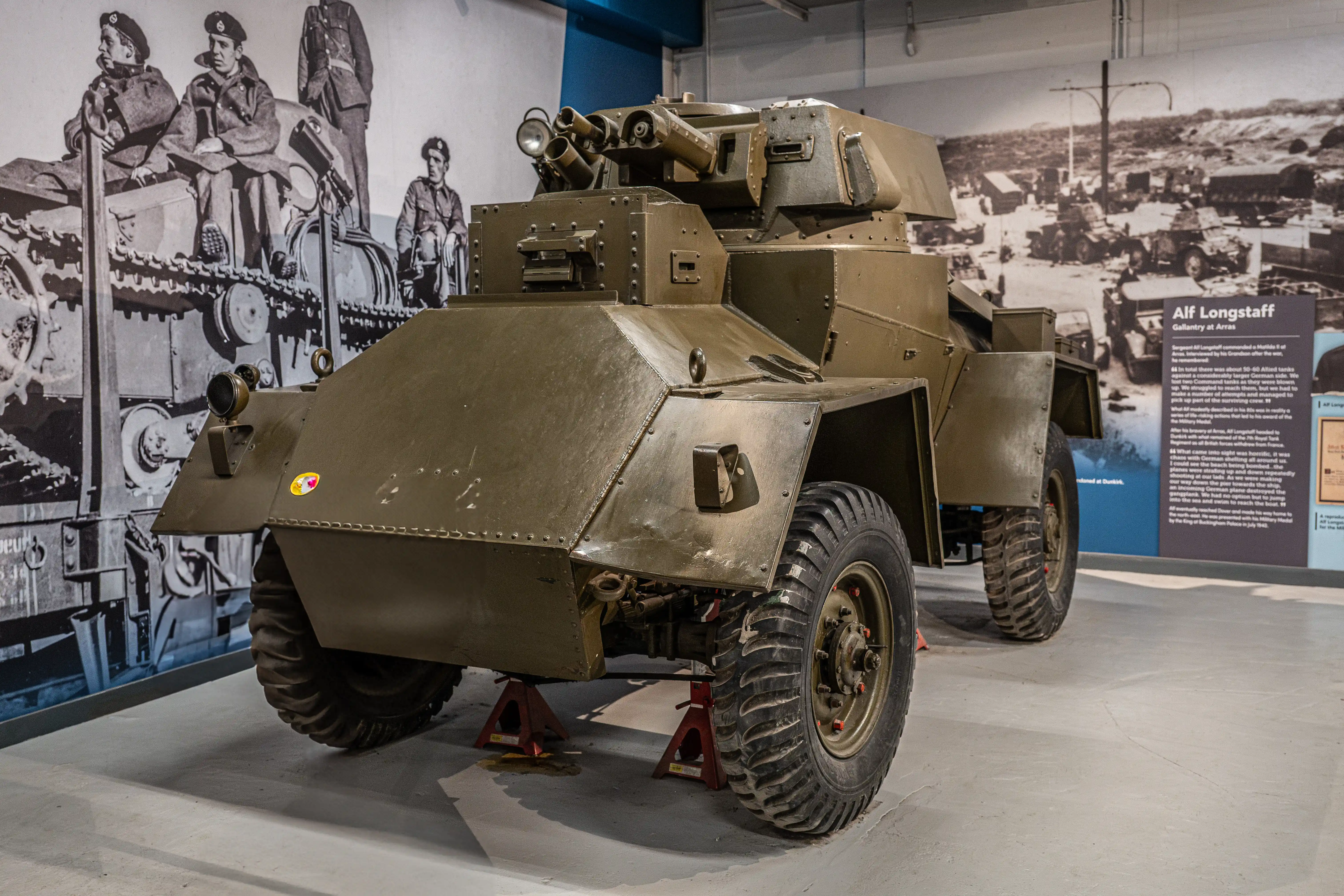 The image depicts a military vehicle, specifically an armored car, displayed in a museum setting. The vehicle is elevated on stands, likely for maintenance or display purposes. Surrounding the vehicle are historical photographs and text panels, one of which highlights the story of Alf Longstaff, a gallantry recipient. The context suggests the vehicle and the displayed information are related to historical military events, possibly from World War II, given the attire of the soldiers in the photographs and the type of vehicle shown.