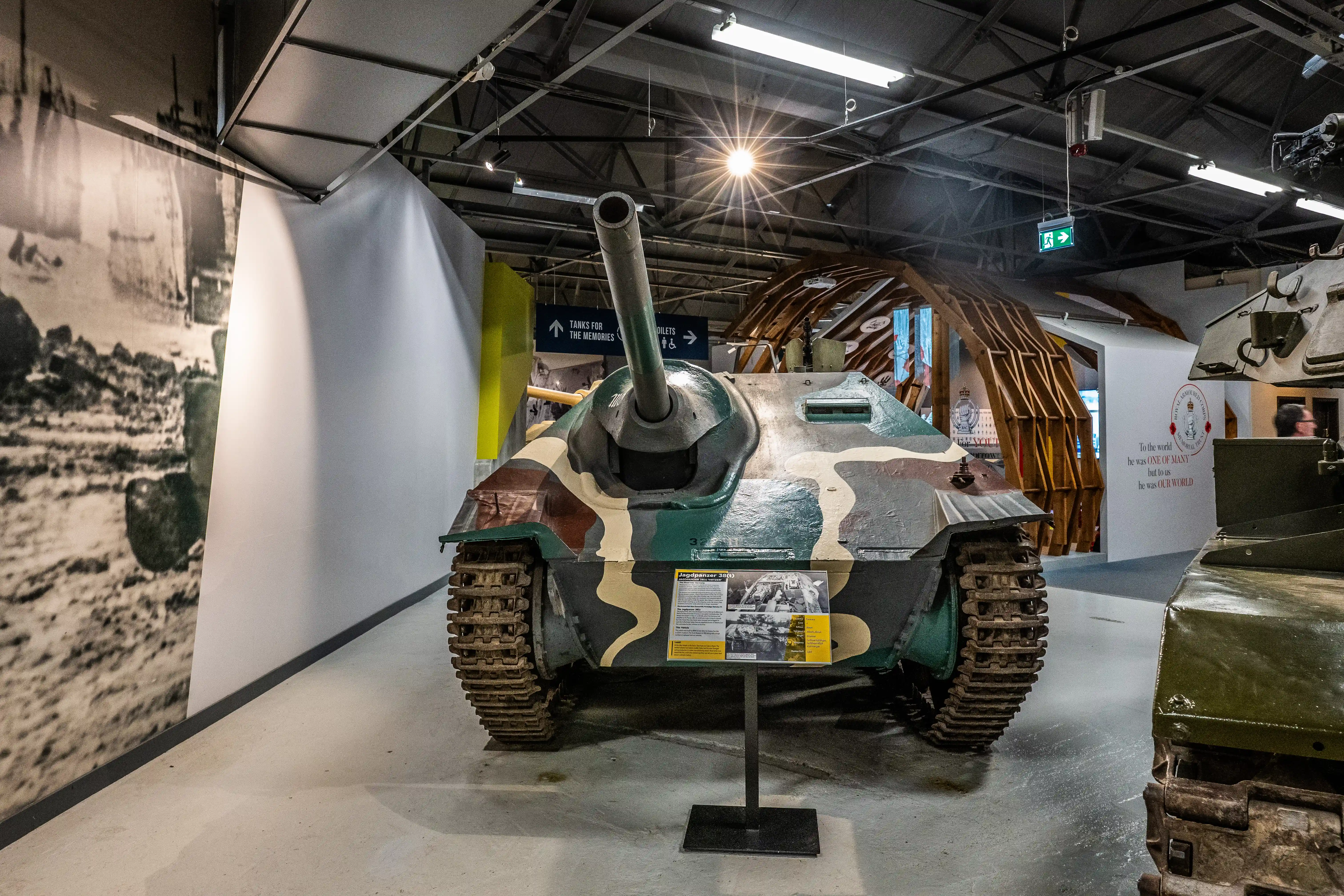 The image depicts a museum exhibit featuring a military tank with camouflage paint. The tank is displayed indoors, with informational placards in front of it. The exhibit is part of a larger display that includes other military vehicles and historical photographs. The setting appears to be well-lit, with a modern, clean design, and there are signs directing visitors to different sections of the exhibit.