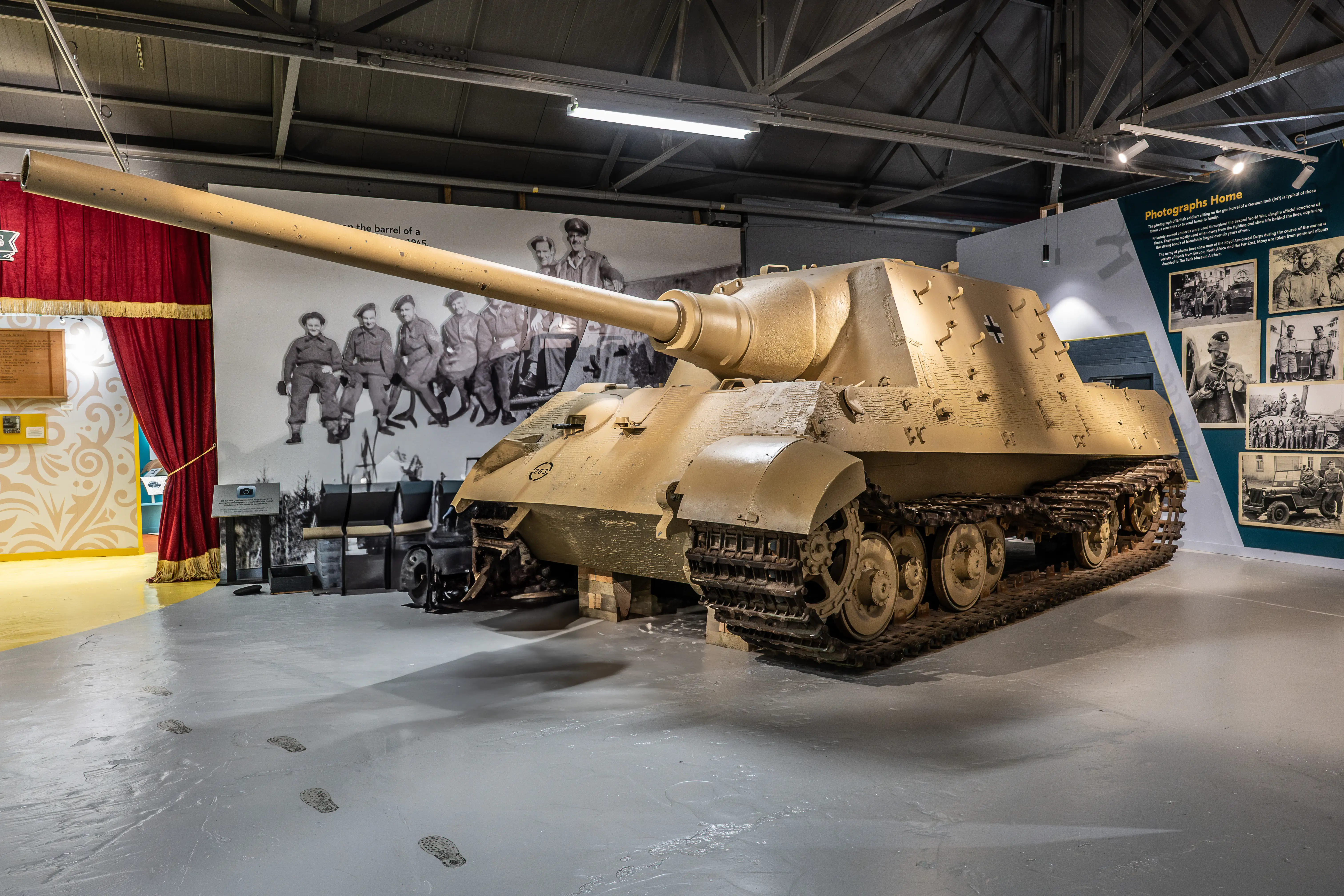 The image depicts a museum exhibit featuring a large, historical military tank. The tank is displayed prominently in an indoor setting, with various informational displays and photographs around it. The photographs show soldiers and other historical images, likely related to the tank's operational history. The setting appears to be well-lit and designed to provide educational context about the tank and its significance.