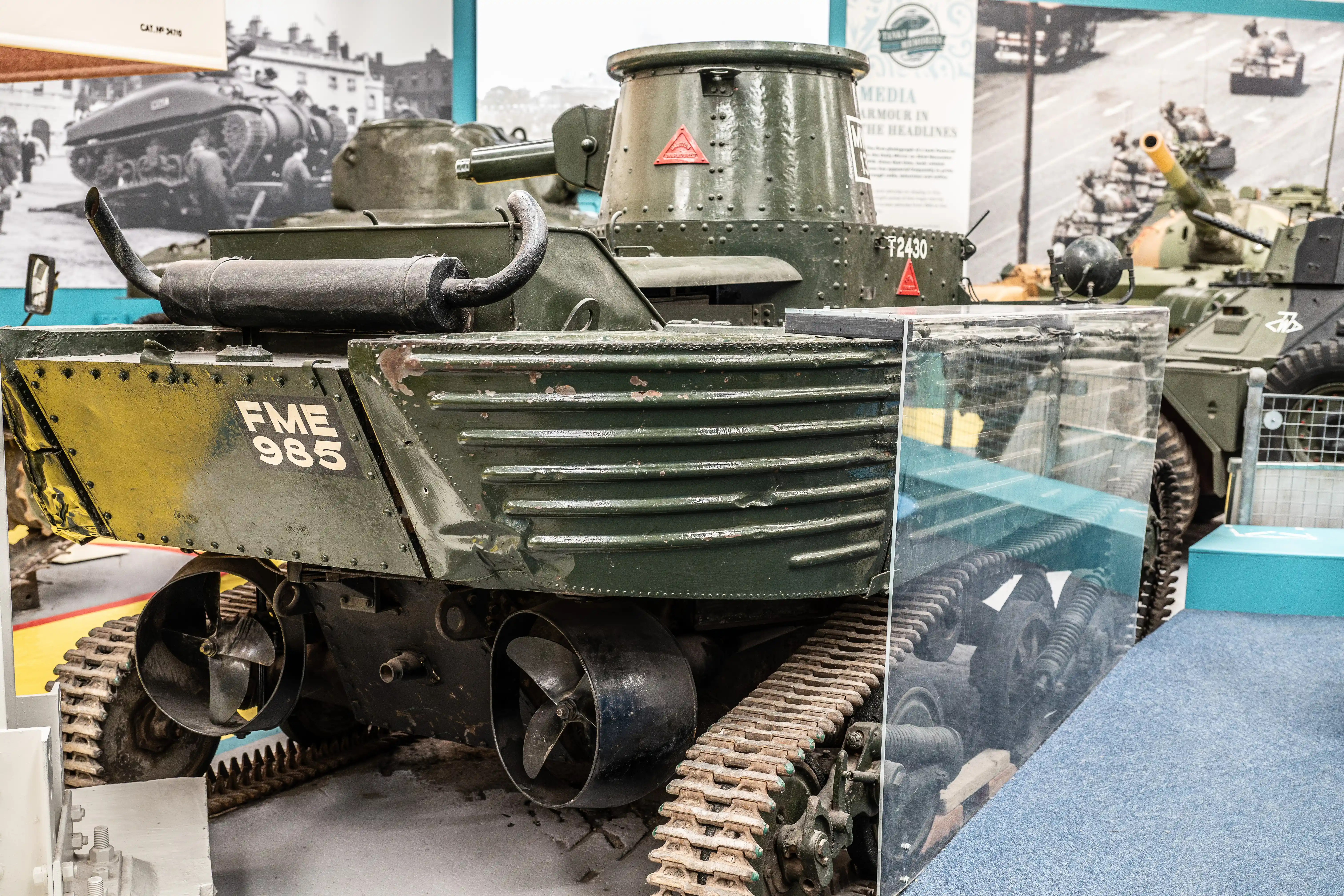 The image depicts a vintage military tank on display, likely in a museum setting. The tank is marked with the identification number FME 985 and features a green and yellow color scheme. The tank's tracks and undercarriage are prominently visible, showcasing detailed components and mechanical parts. Surrounding the tank are informational displays and photographs, providing historical context and additional information about the tank and its history.